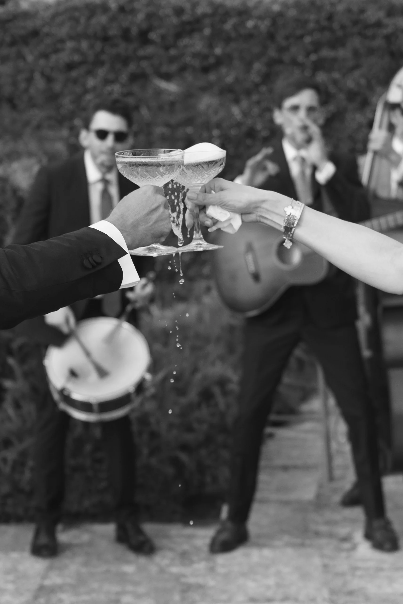 A black-and-white photograph of a wedding toast, with two people clinking champagne glasses. The background shows two musicians, one playing a guitar and the other possibly singing or speaking, with a third instrument resting against a wall.