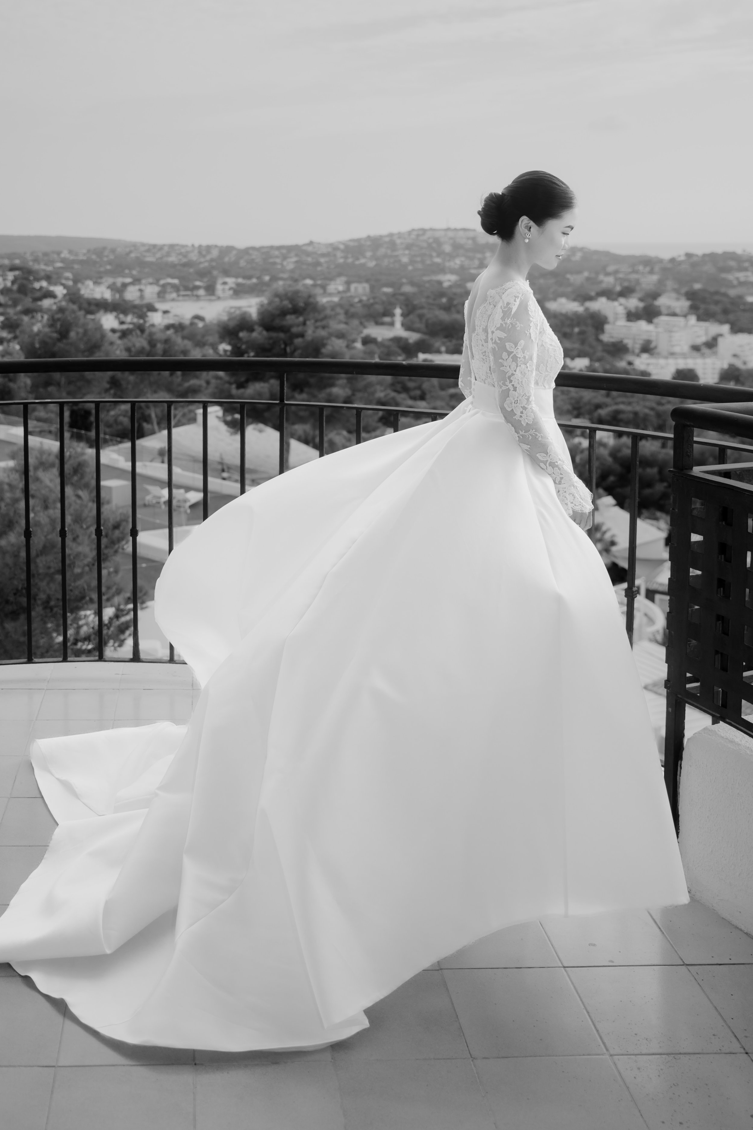 A bride in a white wedding dress with lace sleeves standing on a balcony overlooking a cityscape and hills.