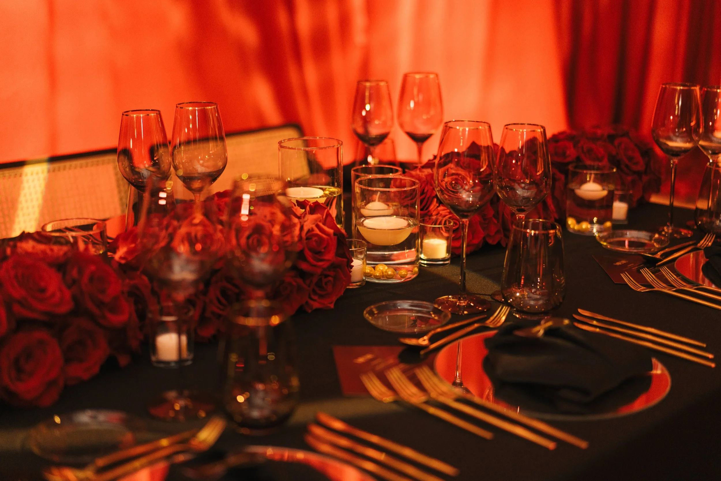 Elegant dining table set with wine glasses, candles, and a floral centerpiece of red roses, illuminated by warm orange lighting.