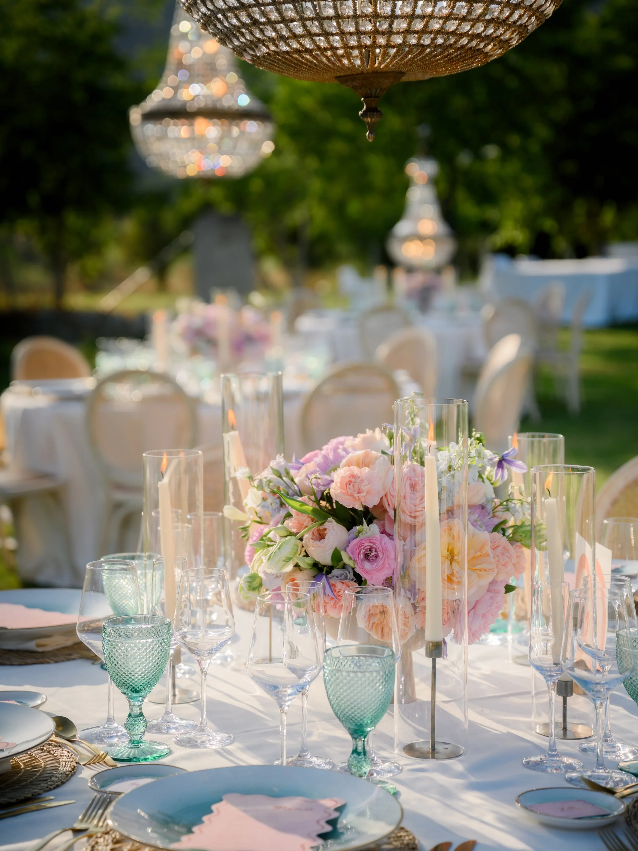 Elegant outdoor banquet table decorated with pink and purple flowers, candles, and glassware, set for a celebration.
