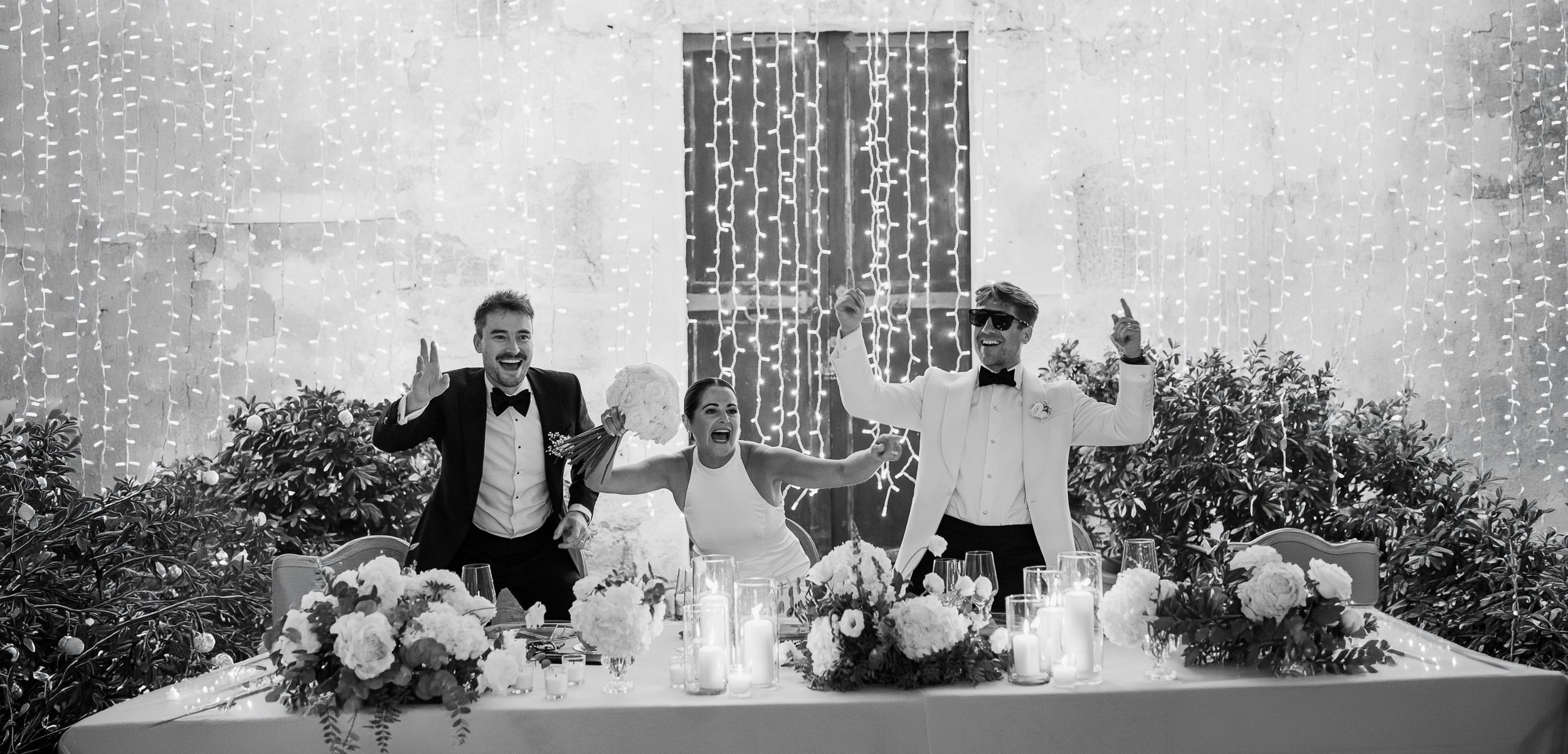A joyful wedding reception scene in black and white, featuring three people at a table with floral centerpieces and candles, with string lights hanging in the background.