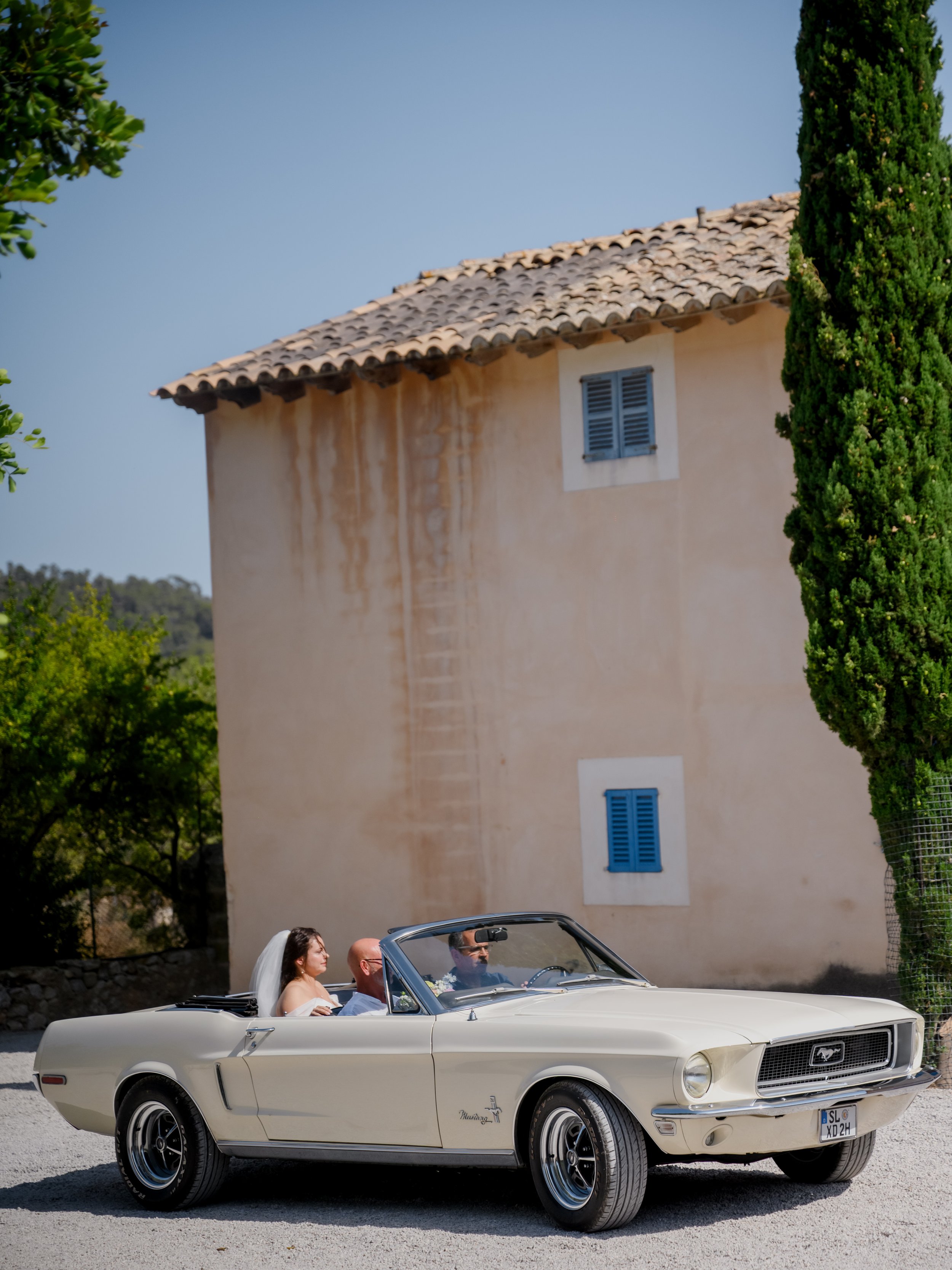 A wedding couple in a white vintage convertible Mustang driving past a beige house with blue shutters.