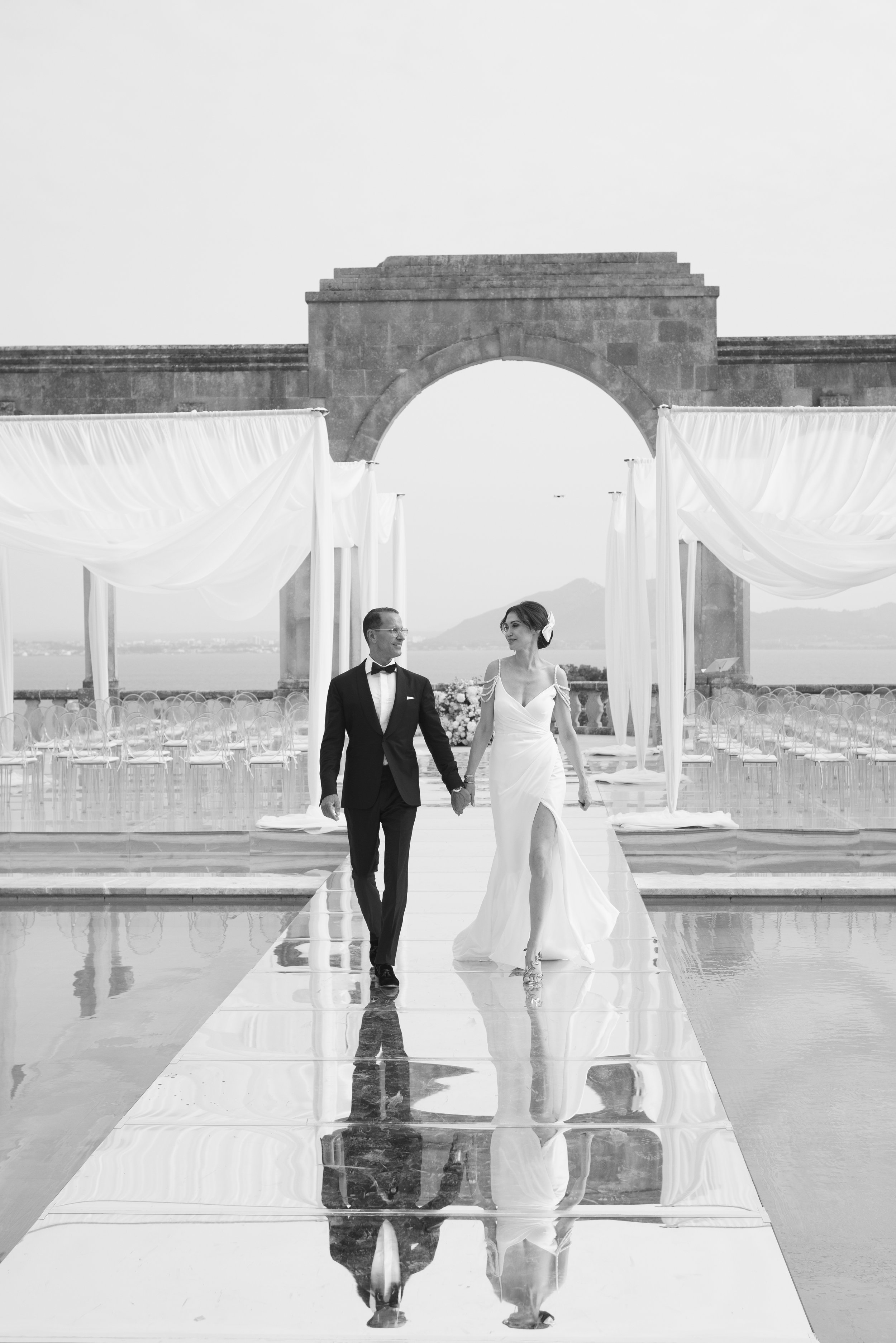 Black and white photo of a bride and groom walking hand in hand down a reflective aisle on an outdoor wedding stage with a mountain and sky backdrop.