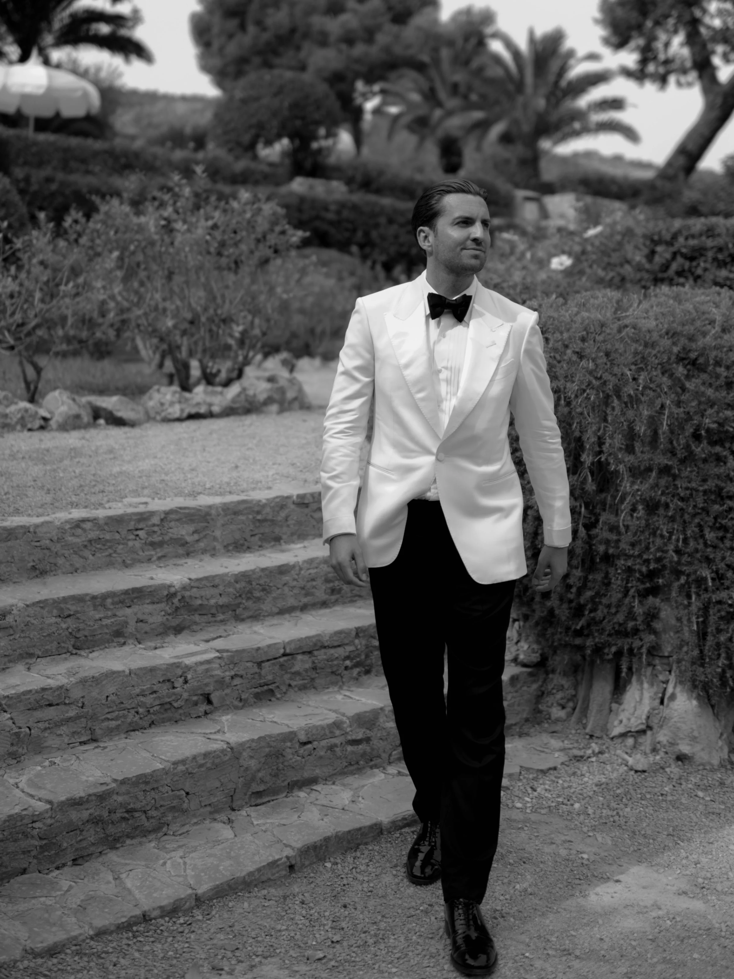 A man wearing formal attire, including a white dinner jacket and black bow tie, walking down stone steps outdoors with bushes and trees in the background.