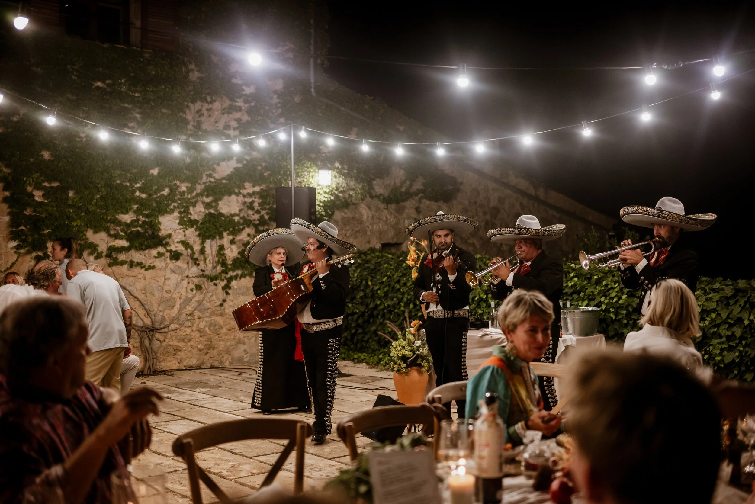 Mariachi band performing at a dinner event outdoors at night under string lights, with guests seated at tables.
