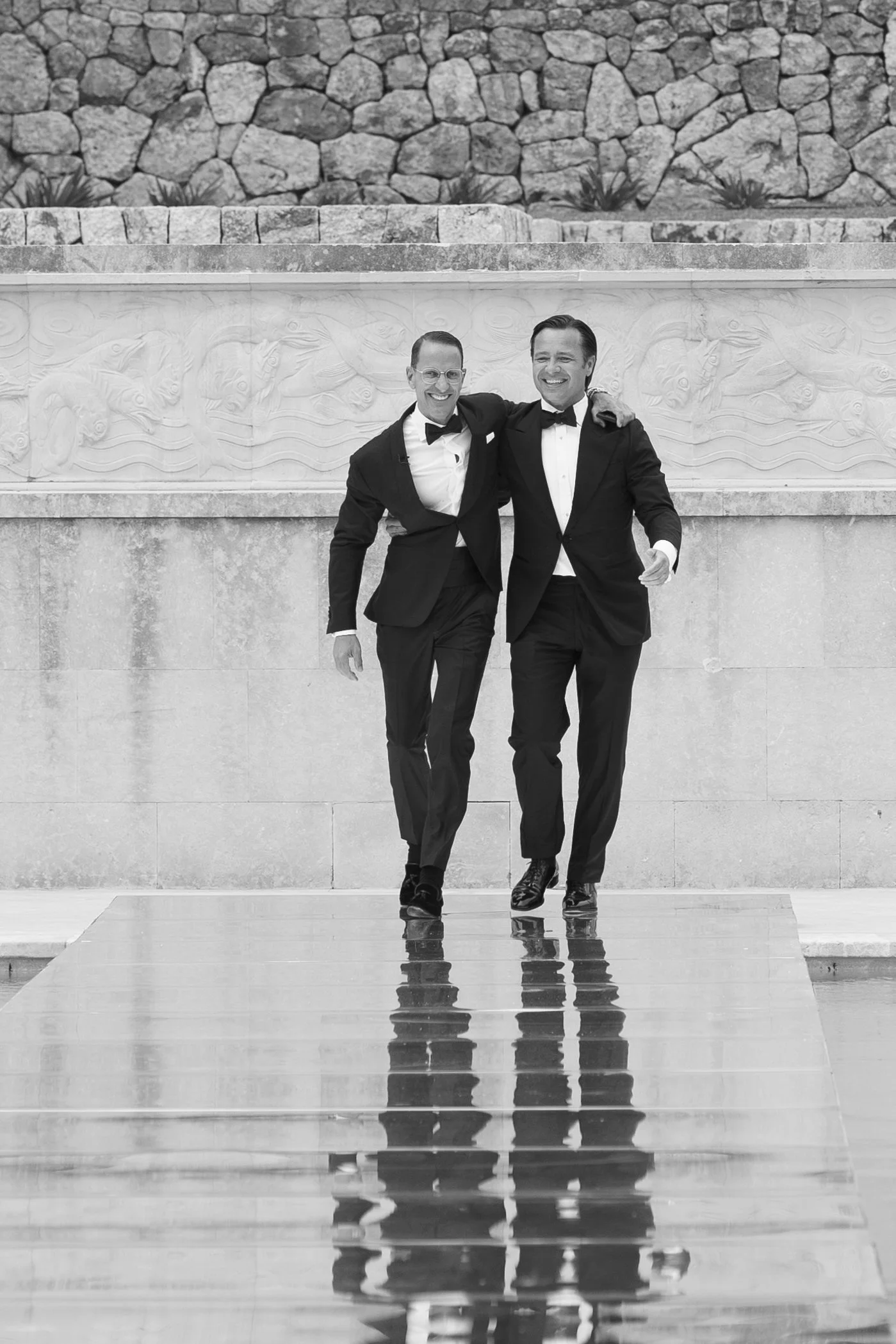 Two men in tuxedos walking arm in arm, smiling, with a stone wall and carved stone panel in the background, and their reflection visible on the shiny floor.