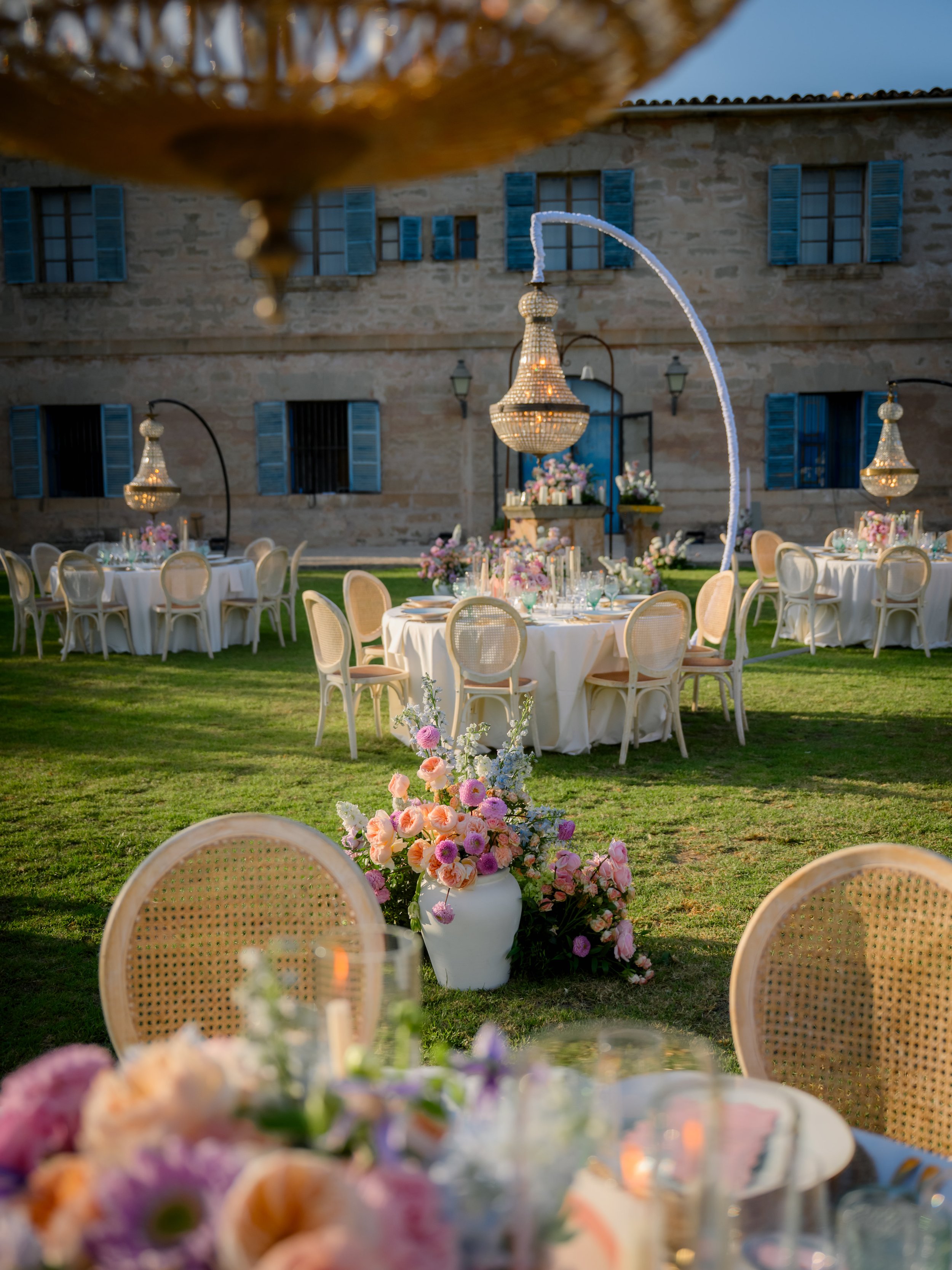 Outdoor wedding reception with decorated tables, floral arrangements, and chandeliers in a garden area in front of a stone building with blue shutters.