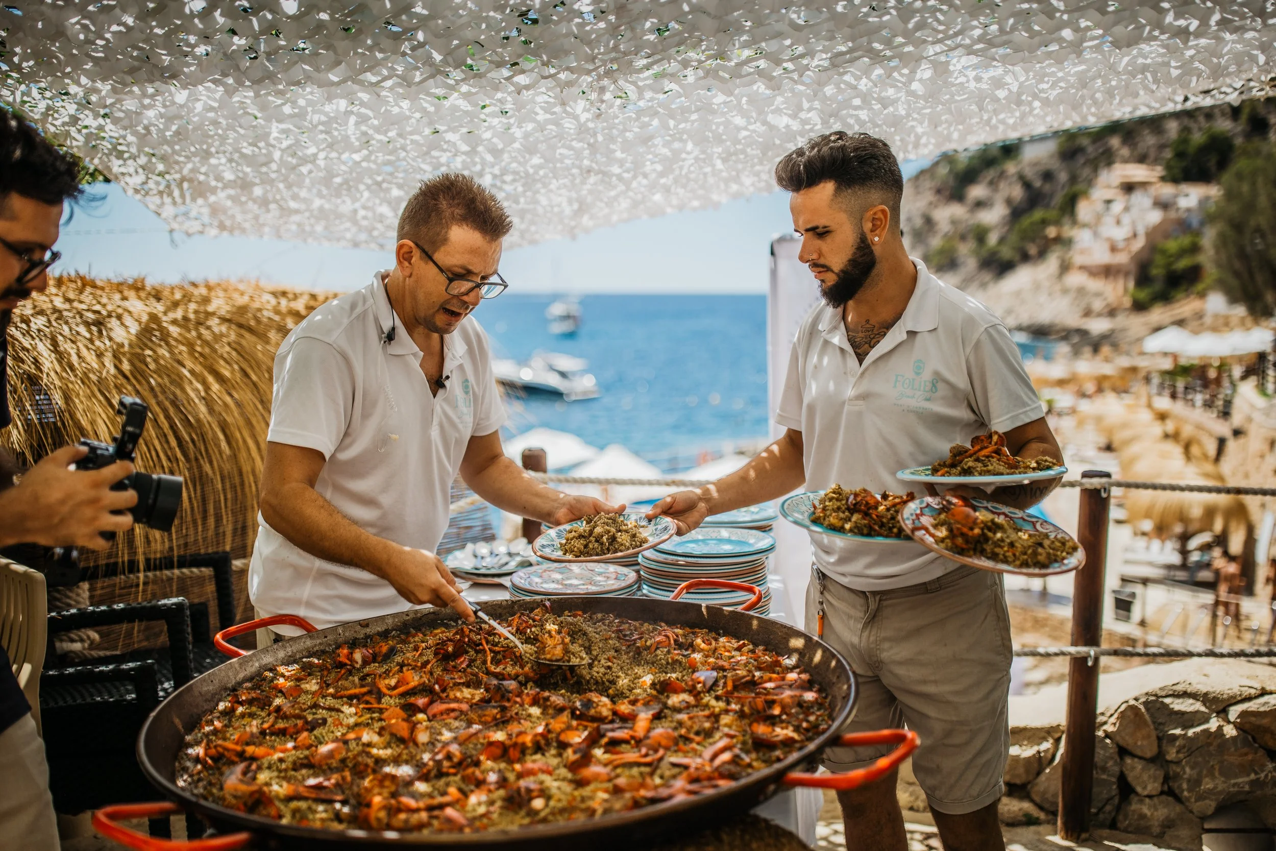 Two men serving seafood paella at an outdoor seaside restaurant with a view of the ocean in the background. One man is holding a plate, and the other is filling it with food.
