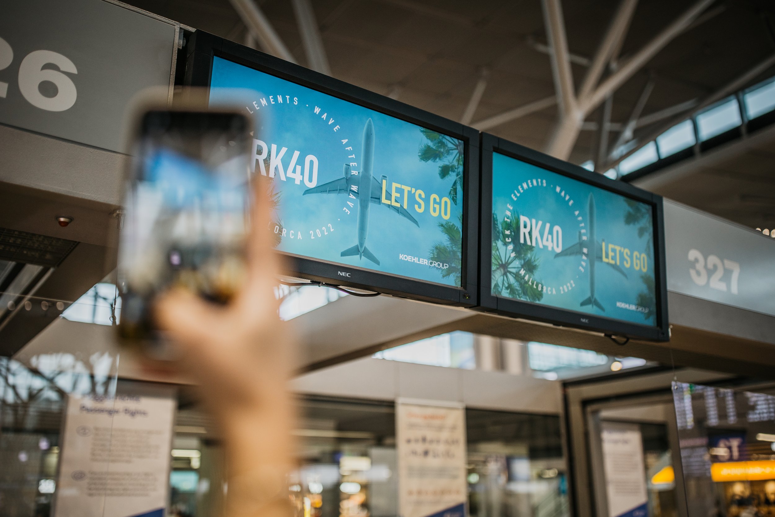 Blurred person holding a phone taking a photo of two digital airport arrival screens showing an advertisement with a plane and the text 'Let's Go' and 'RK40'.