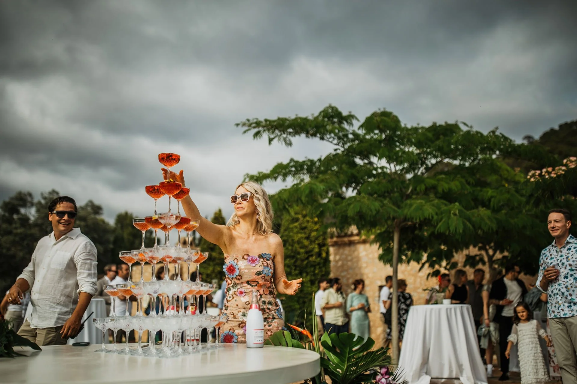 Woman in floral dress stacking glasses to form a champagne tower at an outdoor gathering, with people and trees in the background under cloudy sky.