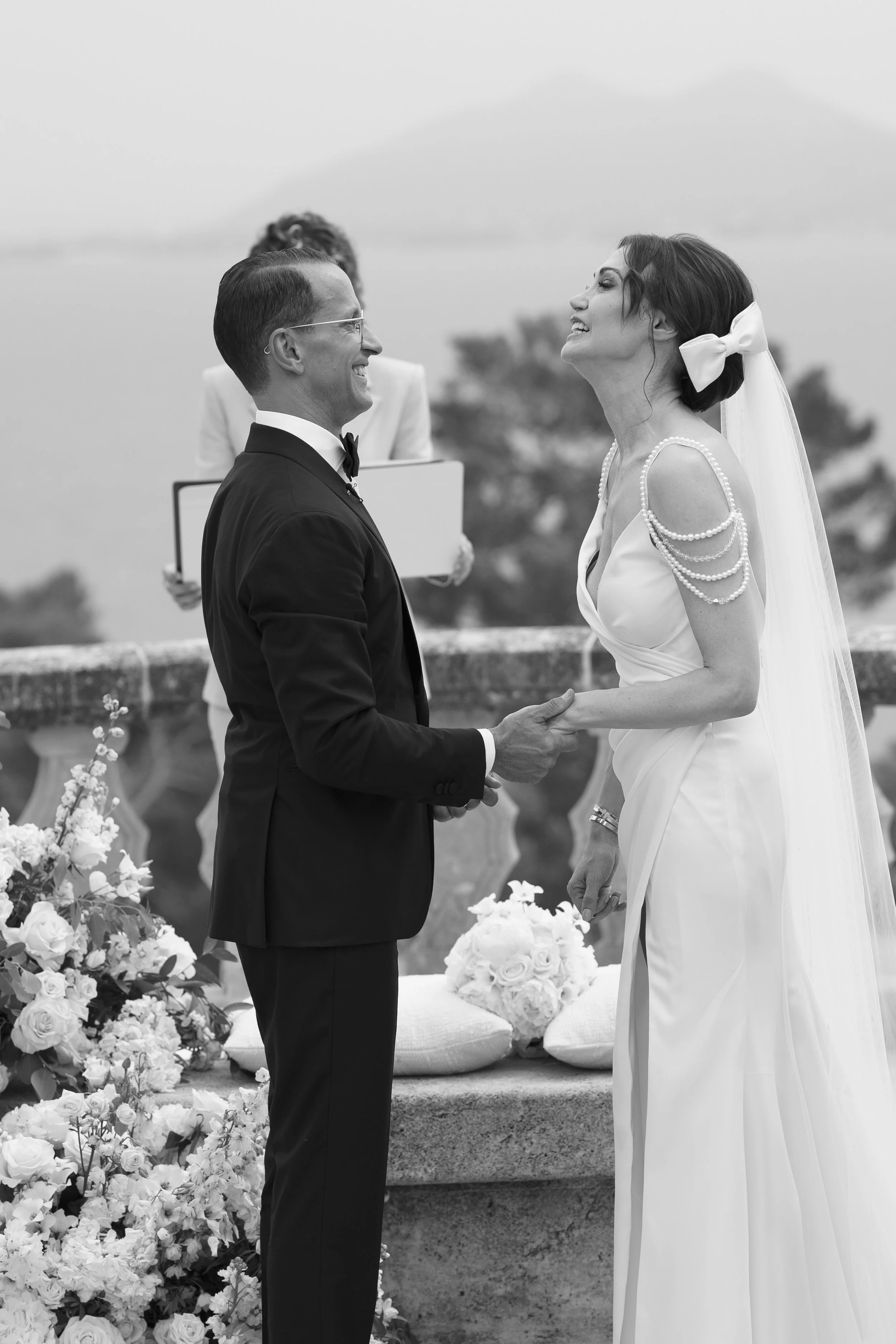 A black and white photo of a wedding ceremony with a bride and groom holding hands and smiling at each other, outdoors with flowers and an officiant in the background.