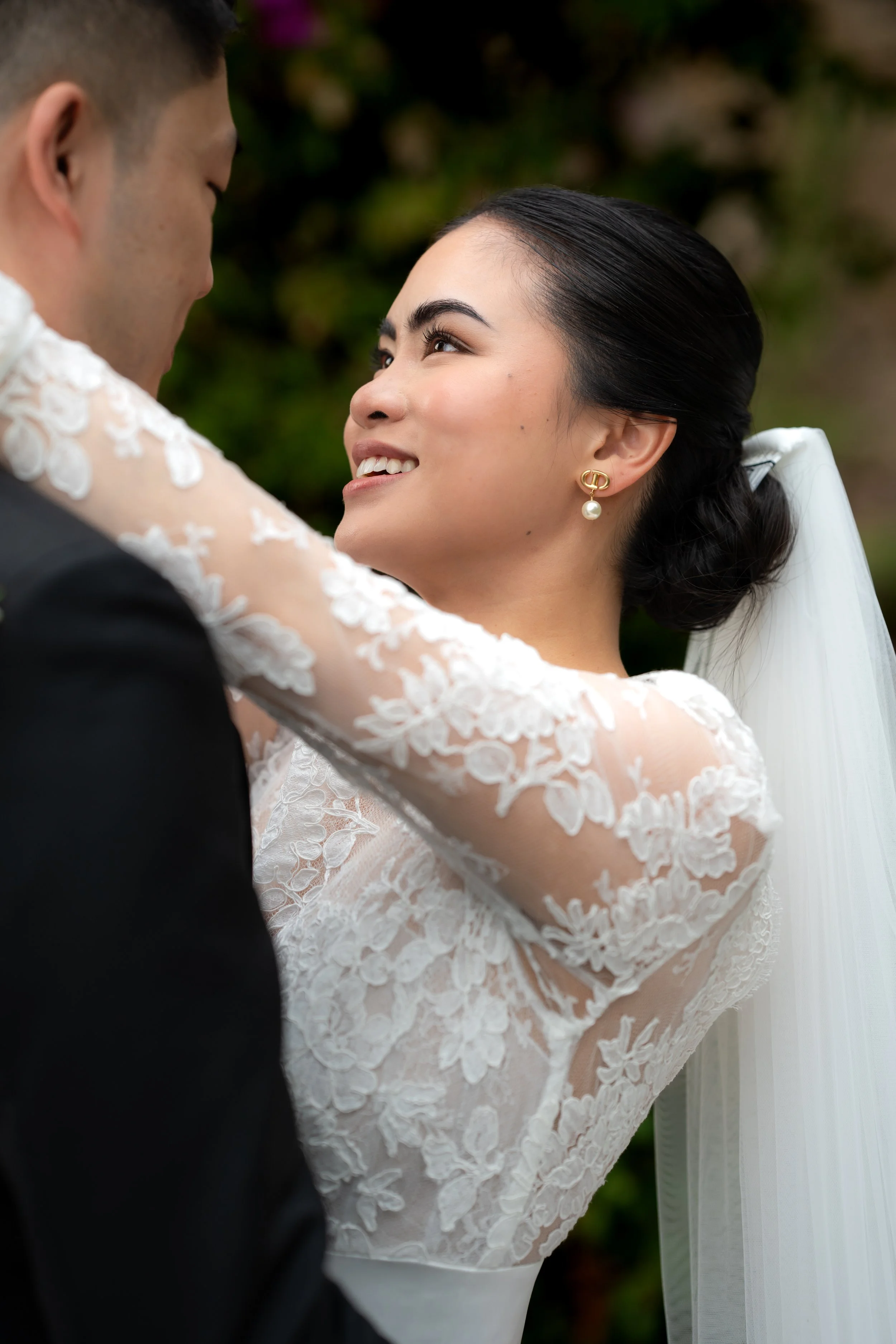 A bride and groom embrace during a wedding ceremony, with the bride smiling and looking into the groom's eyes. The bride is wearing a lace wedding dress with a veil, and has dark hair styled in an elegant updo. The groom is wearing a black suit.