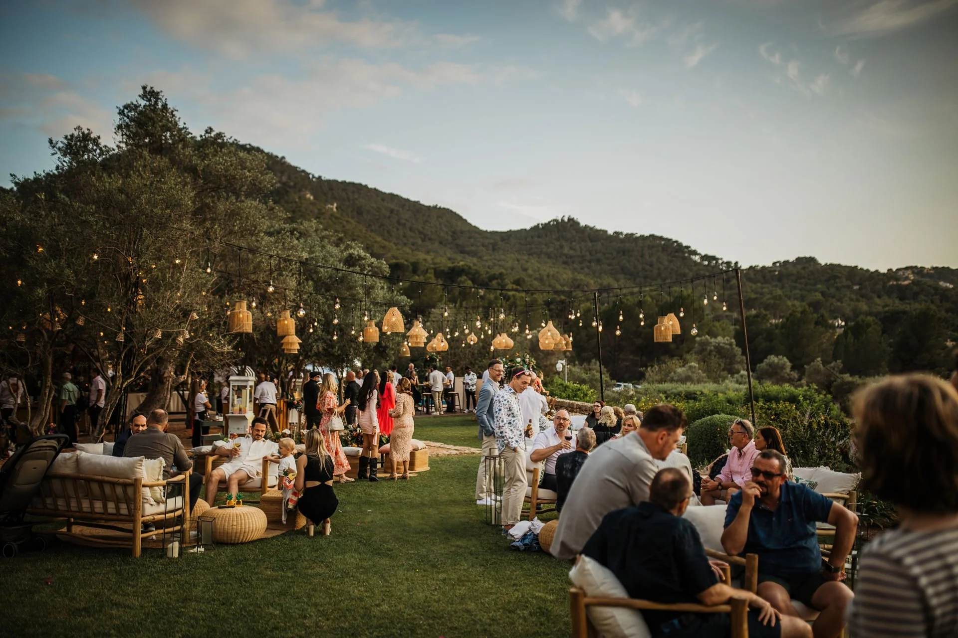 People gathering at an outdoor party or wedding reception in a scenic mountain setting during sunset, with string lights and hanging lanterns.