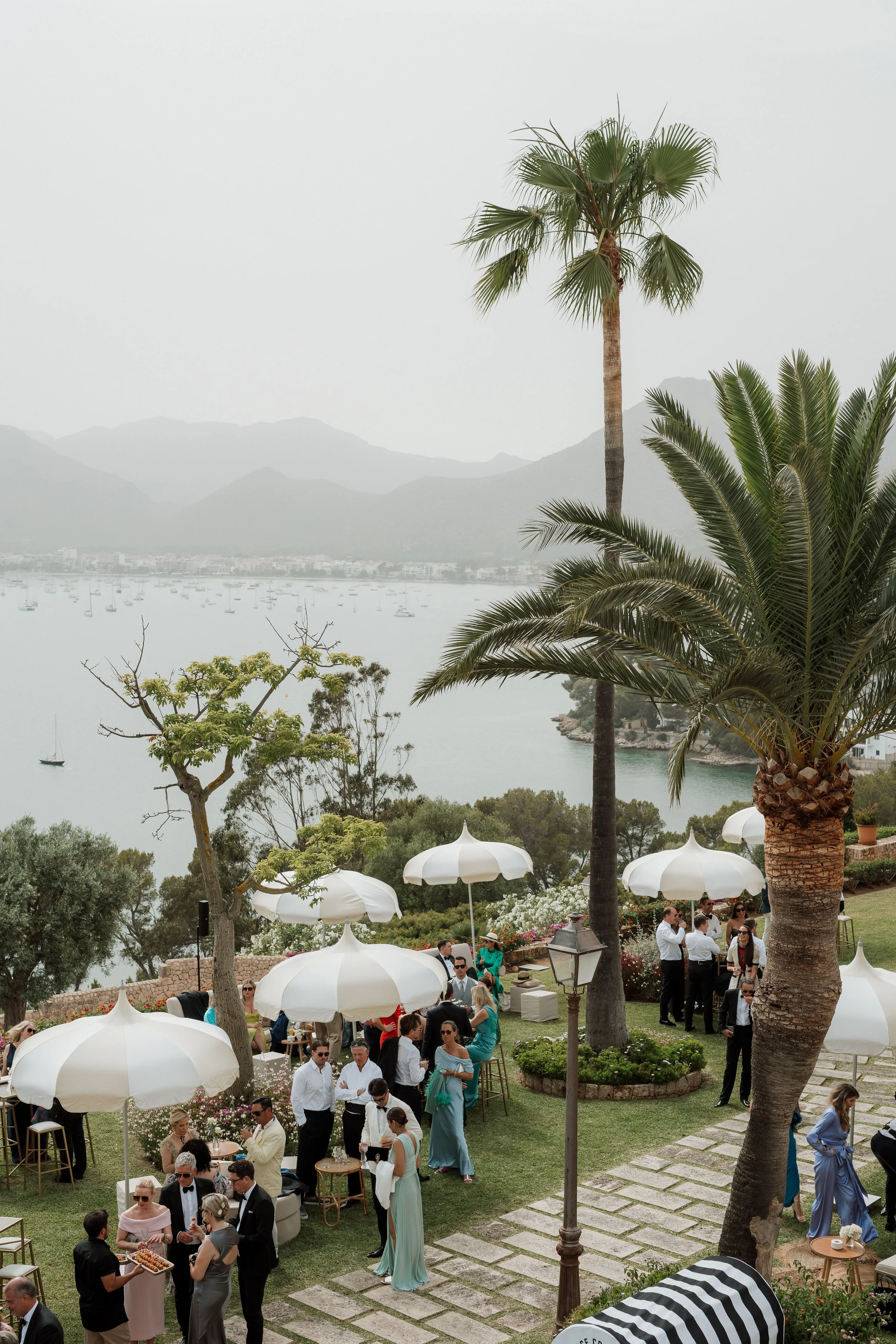 Outdoor social gathering by the water with people, white umbrellas, palm trees, and a scenic mountain lake background.