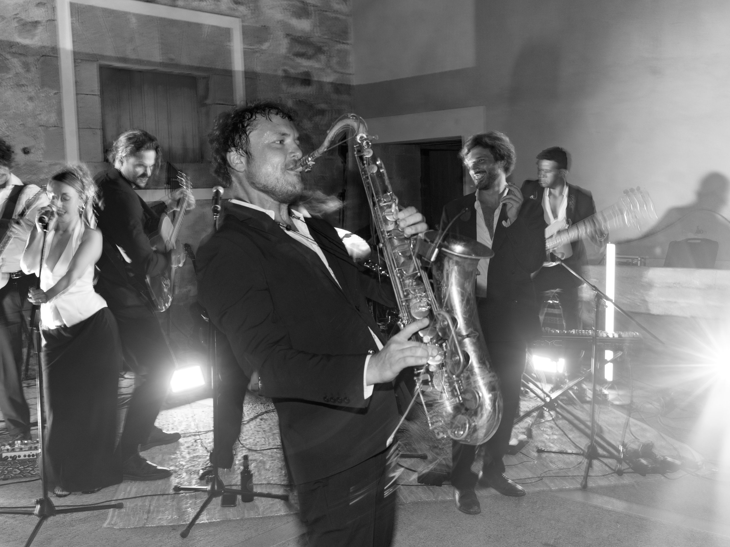 Musicians performing live, with a saxophonist in the foreground and others playing guitar and drums in the background, in a dimly lit indoor venue.