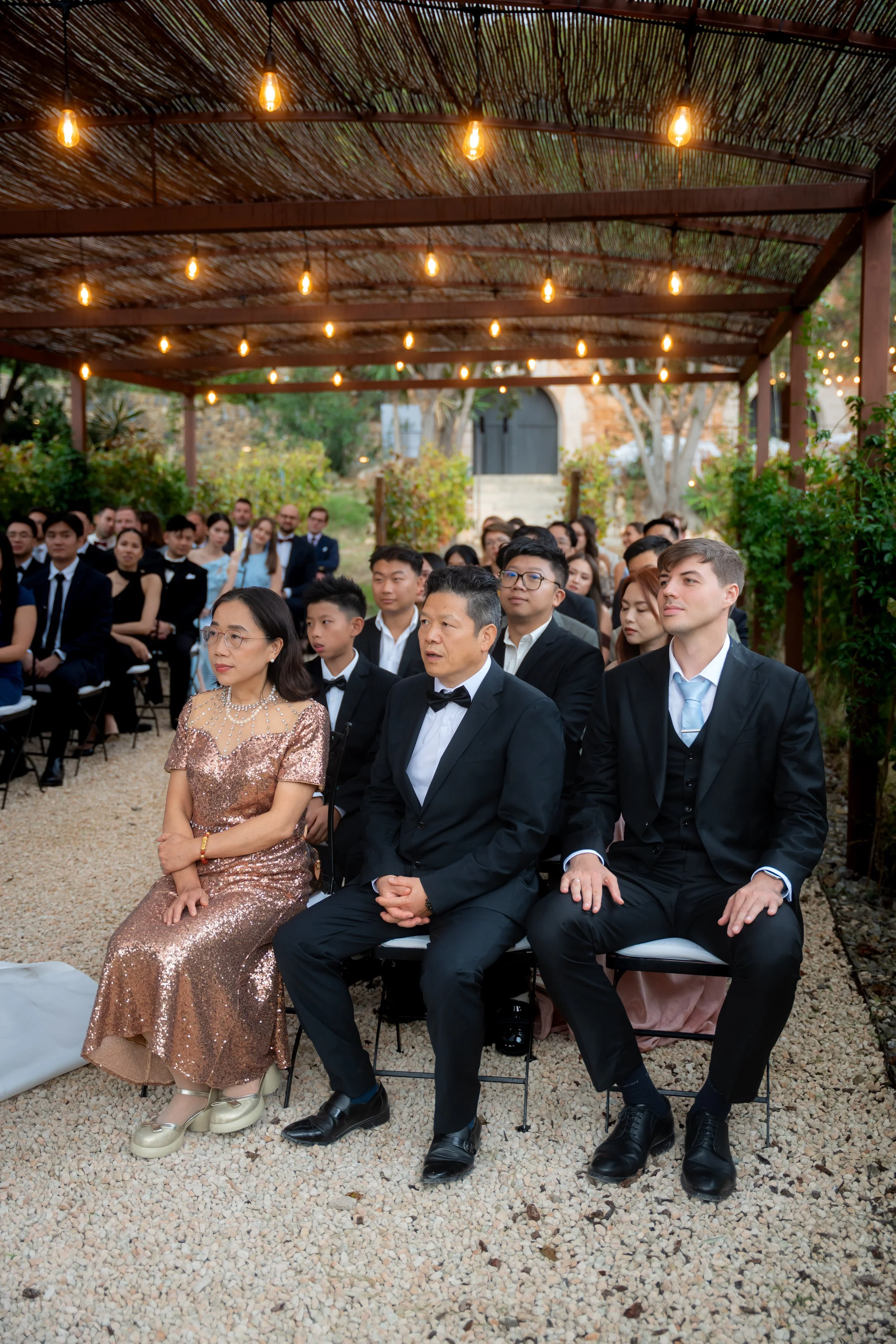 Group of people attending an outdoor wedding ceremony, seated under a wooden pergola decorated with string lights, with greenery and trees in the background.