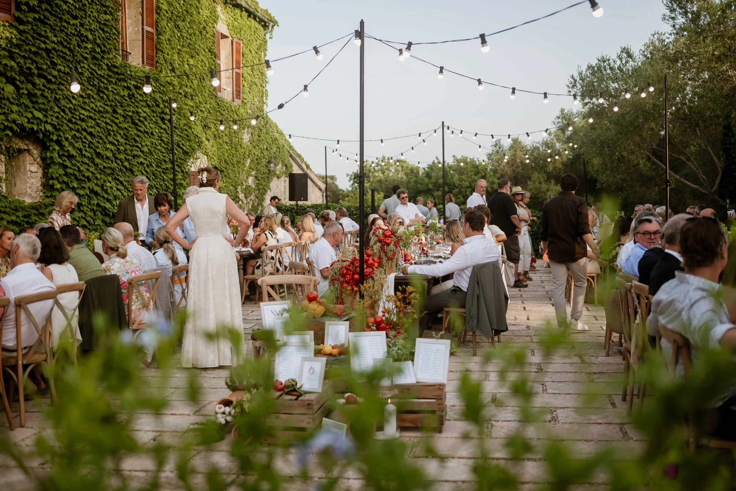 Outdoor wedding reception with guests seated at decorated tables under string lights, surrounded by greenery and ivy-covered walls.