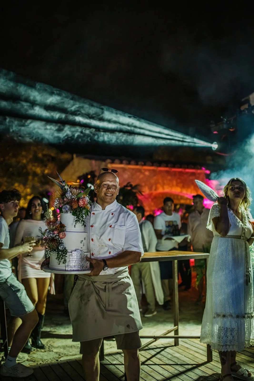 Chef holding a decorated cake at an outdoor party at night with colorful lights and people in the background.
