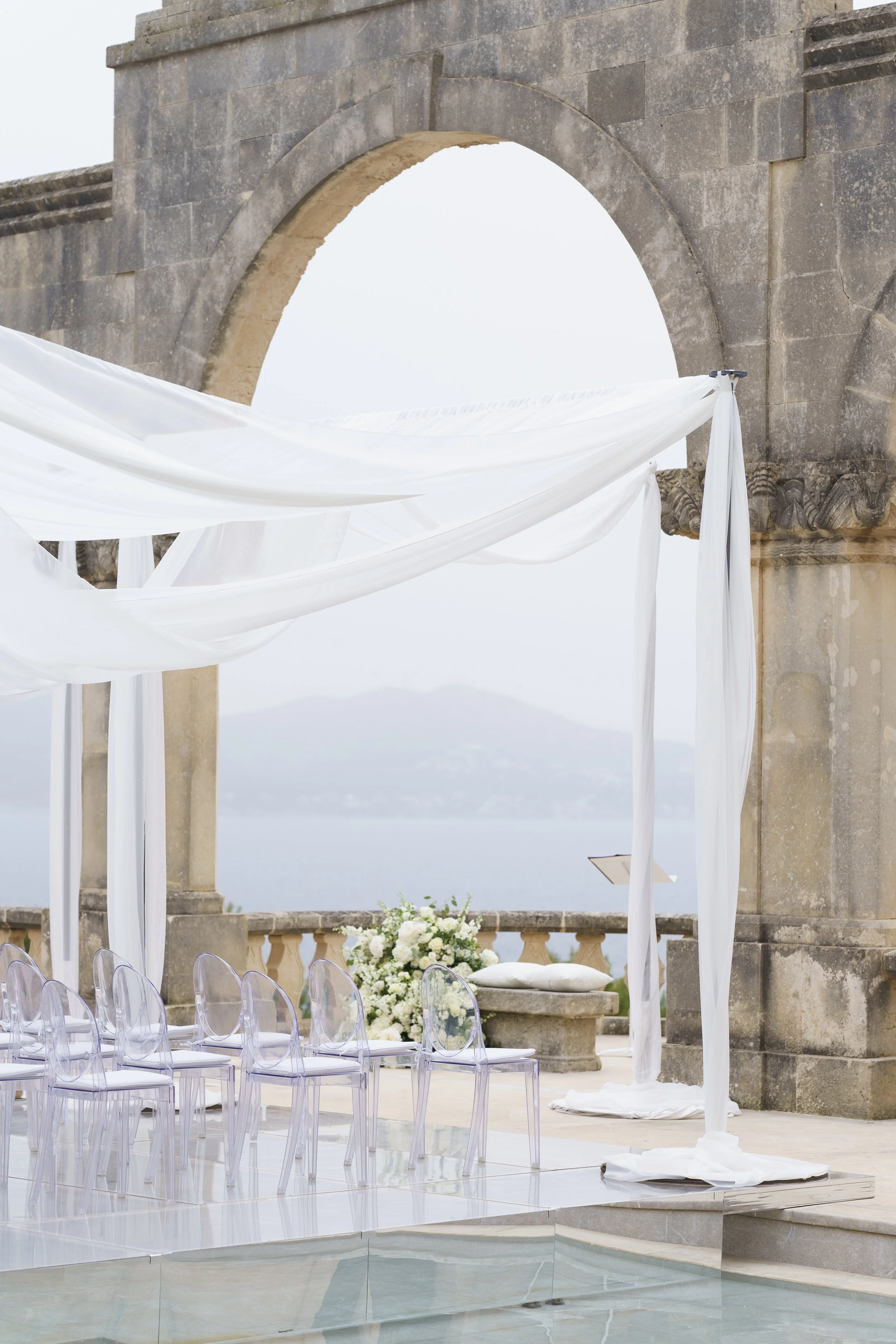 Elegant outdoor wedding altar with white flowing fabric, transparent chairs, floral arrangements, stone arch, overlooking a body of water and distant mountains.