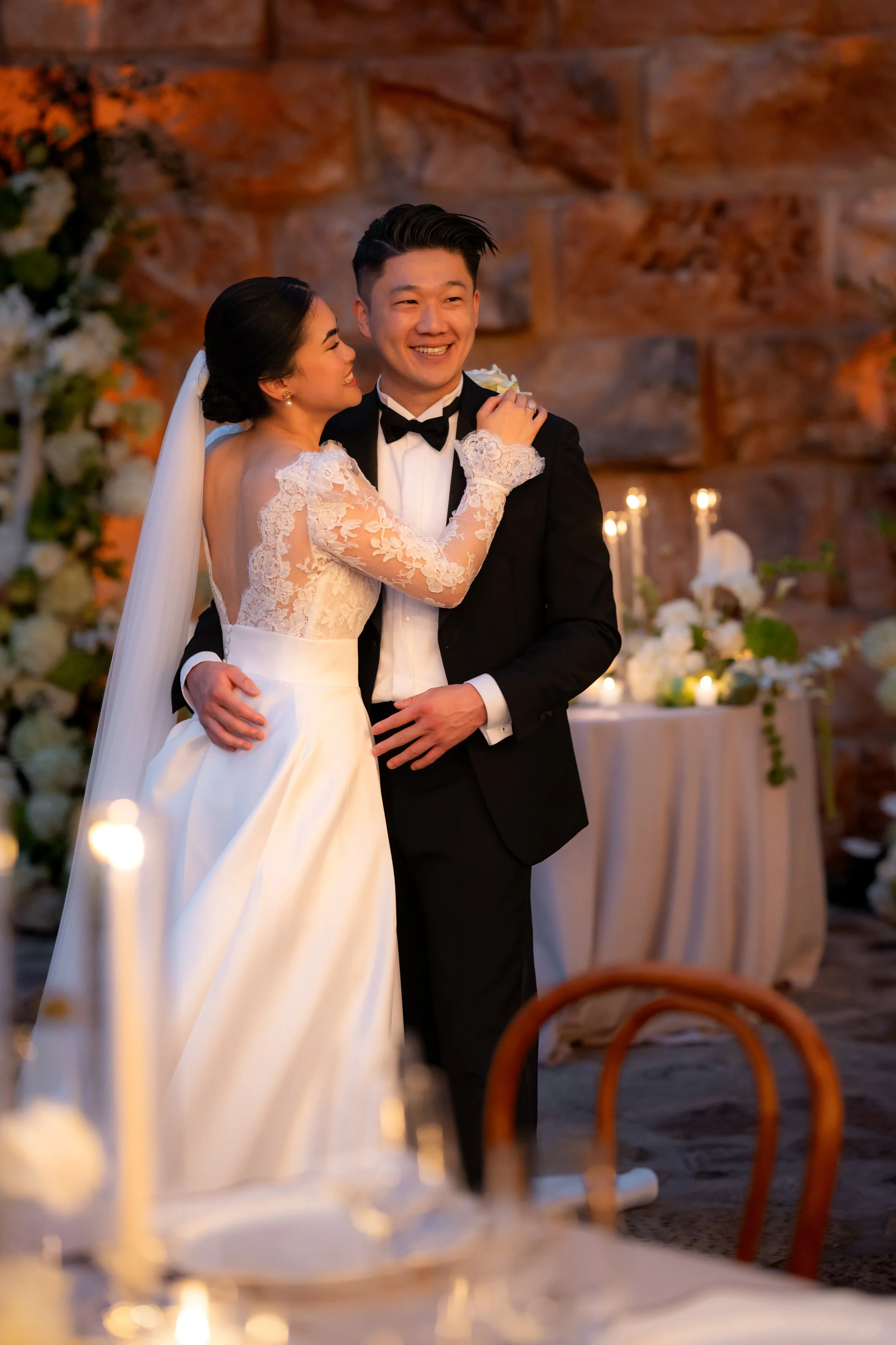 A bride and groom dancing at their wedding reception, smiling and embracing each other, with candlelit decorations in the background.