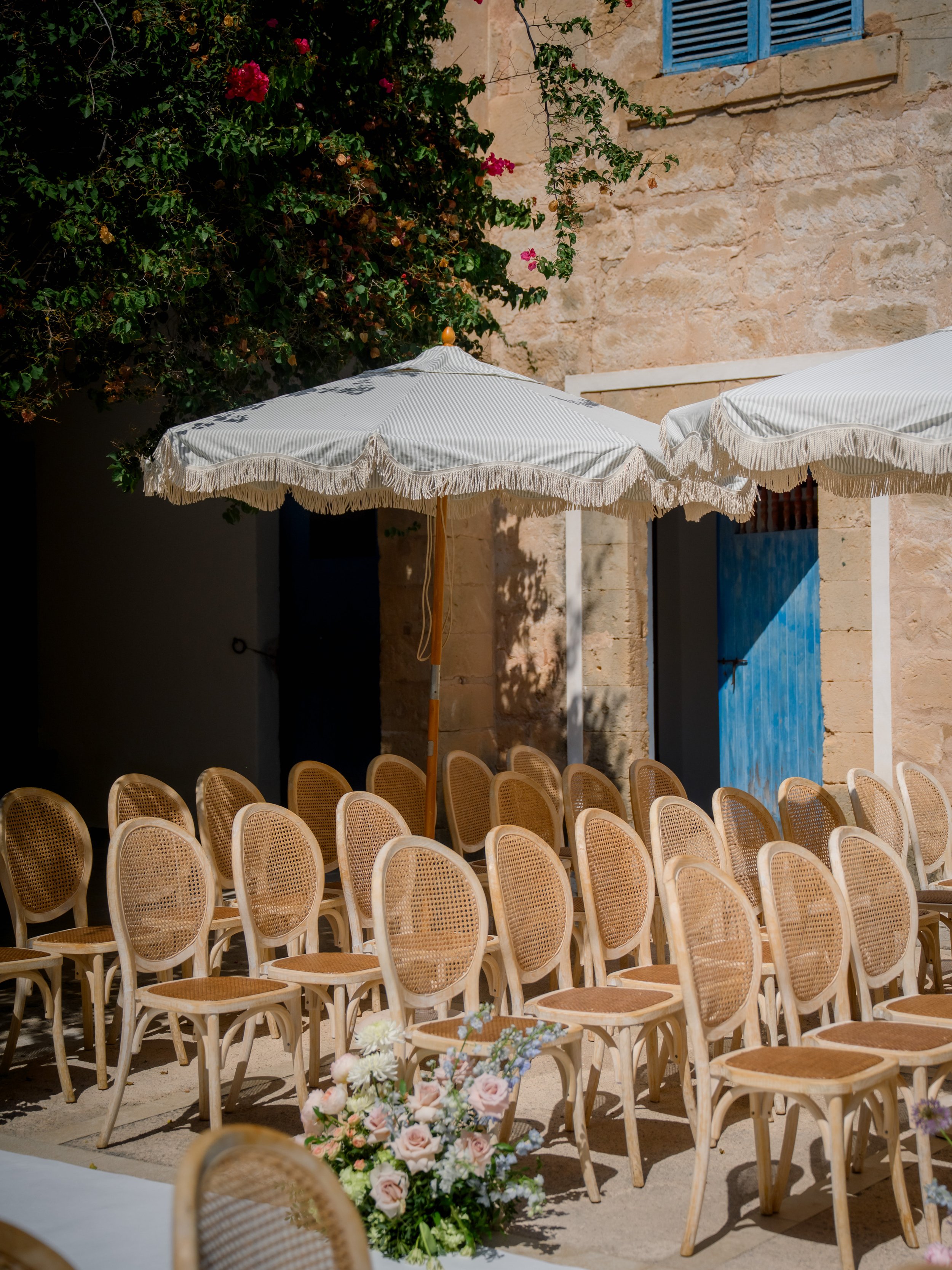 Empty outdoor wedding ceremony setup with rows of chairs, large umbrellas, and floral arrangements on a sunny day.