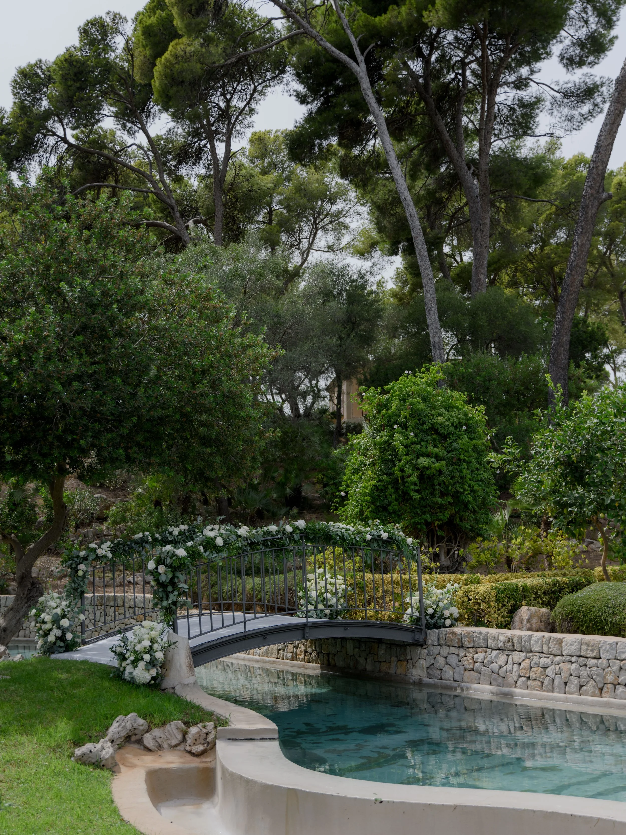A garden with a small bridge decorated with white flowers over a narrow pond, surrounded by green bushes and tall trees.