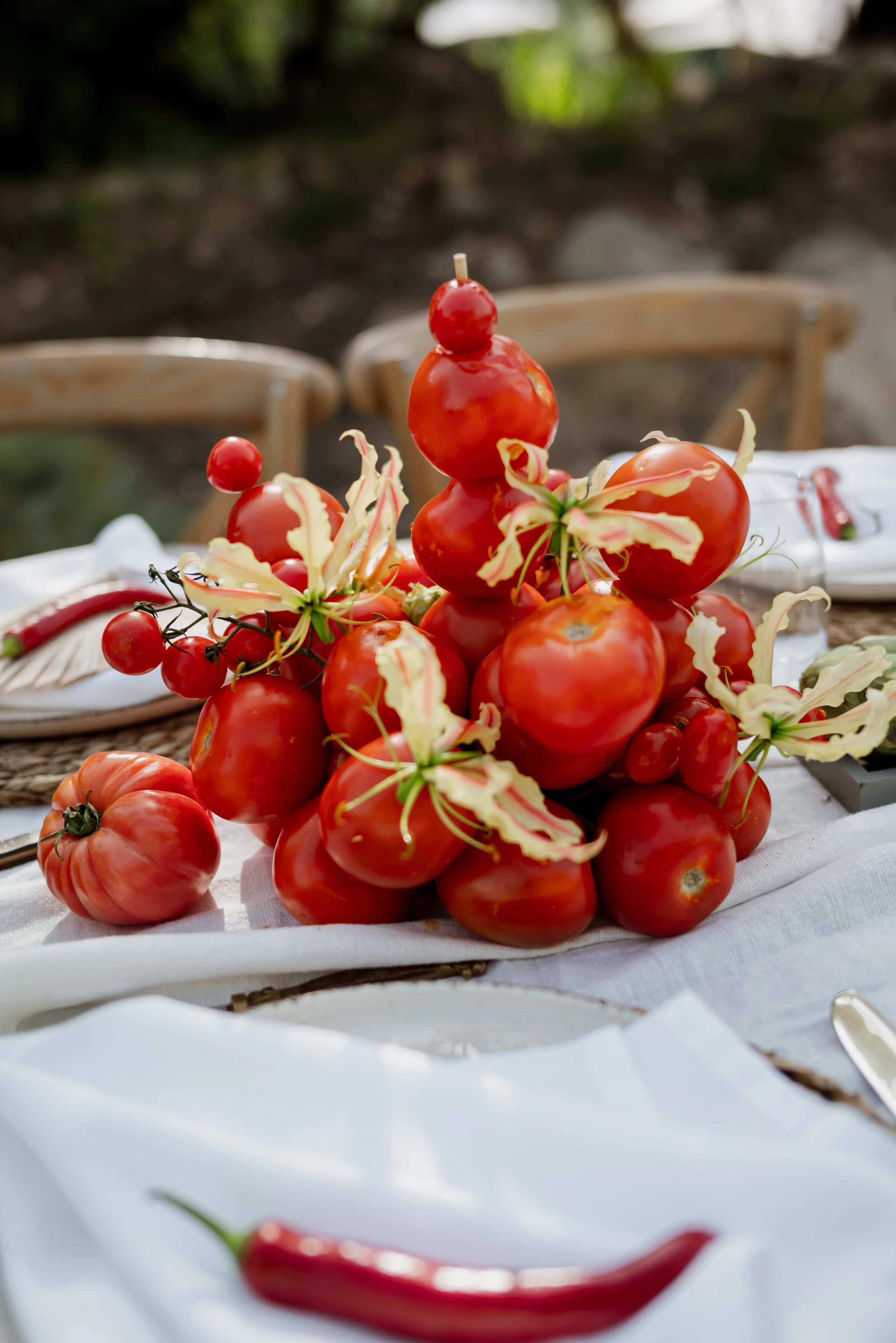 A table decorated with a pile of tomatoes and cherry tomatoes, accented by some flowering branches, set outdoors.