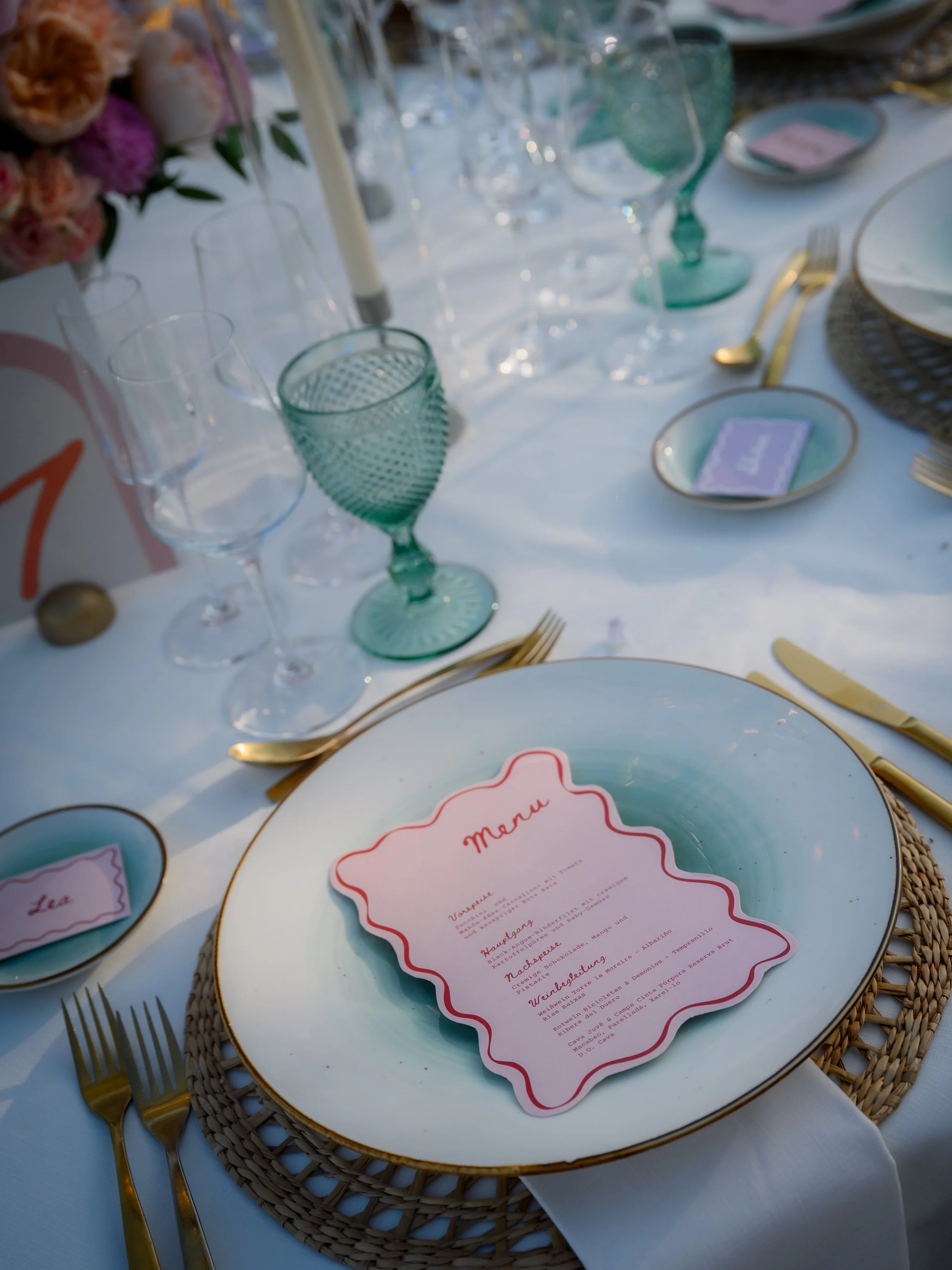 Elegant dinner table setup with a menu on a large white plate, surrounded by gold flatware, various glasses, and small place cards, with a floral centerpiece in the background.