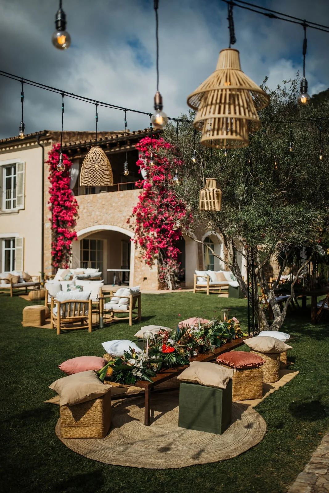 Outdoor event setup with wooden seats, cushions, floral arrangements, and hanging wicker lamps in a garden yard of a house with pink flowering plants.