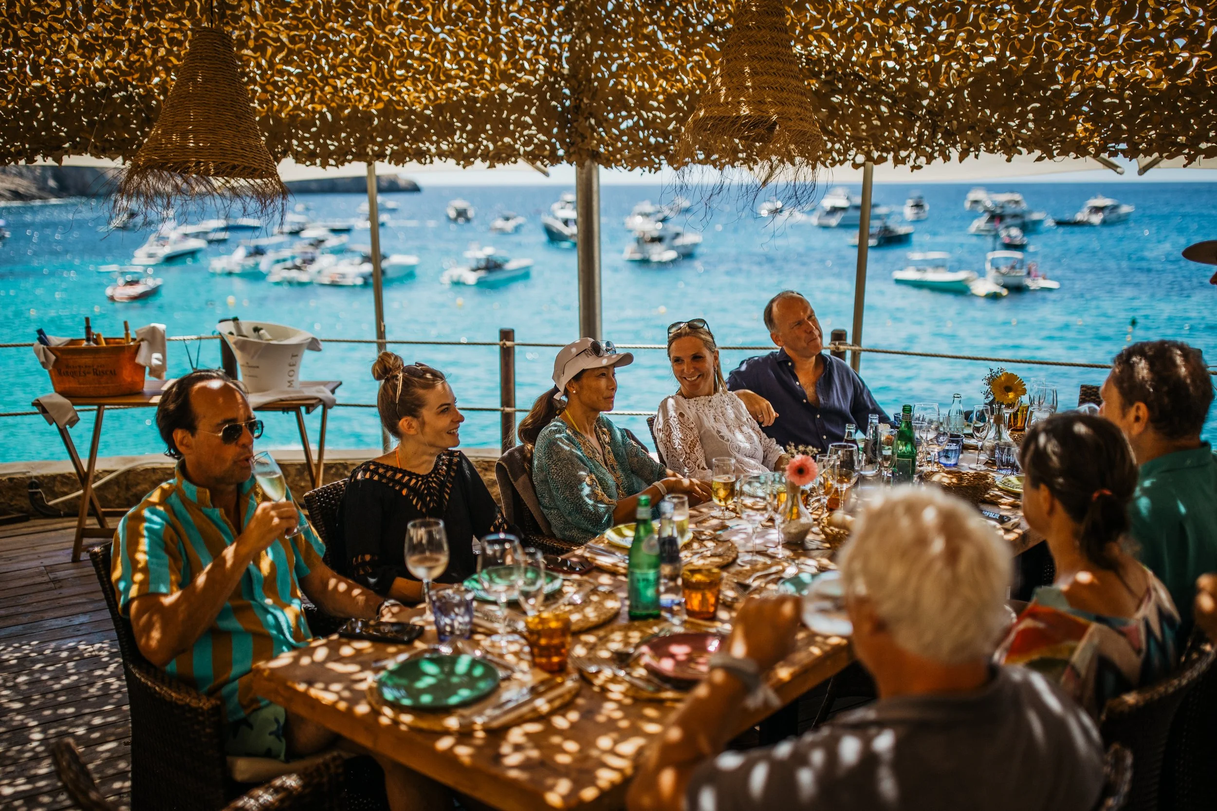 Group of people dining at an outdoor restaurant overlooking the ocean, with boats anchored in the water.