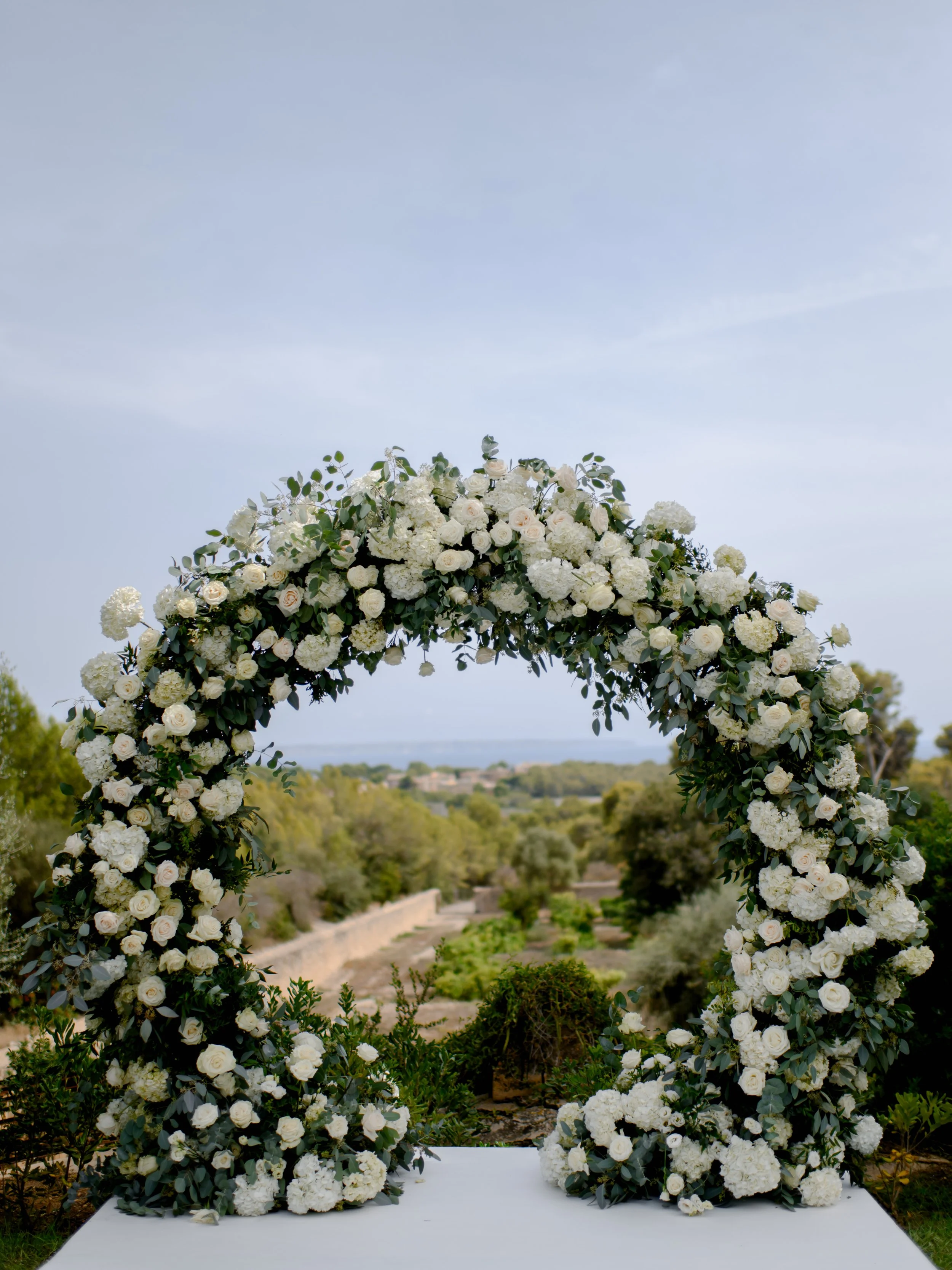 A floral arch decorated with white roses and hydrangeas, set outdoors with a scenic landscape background.