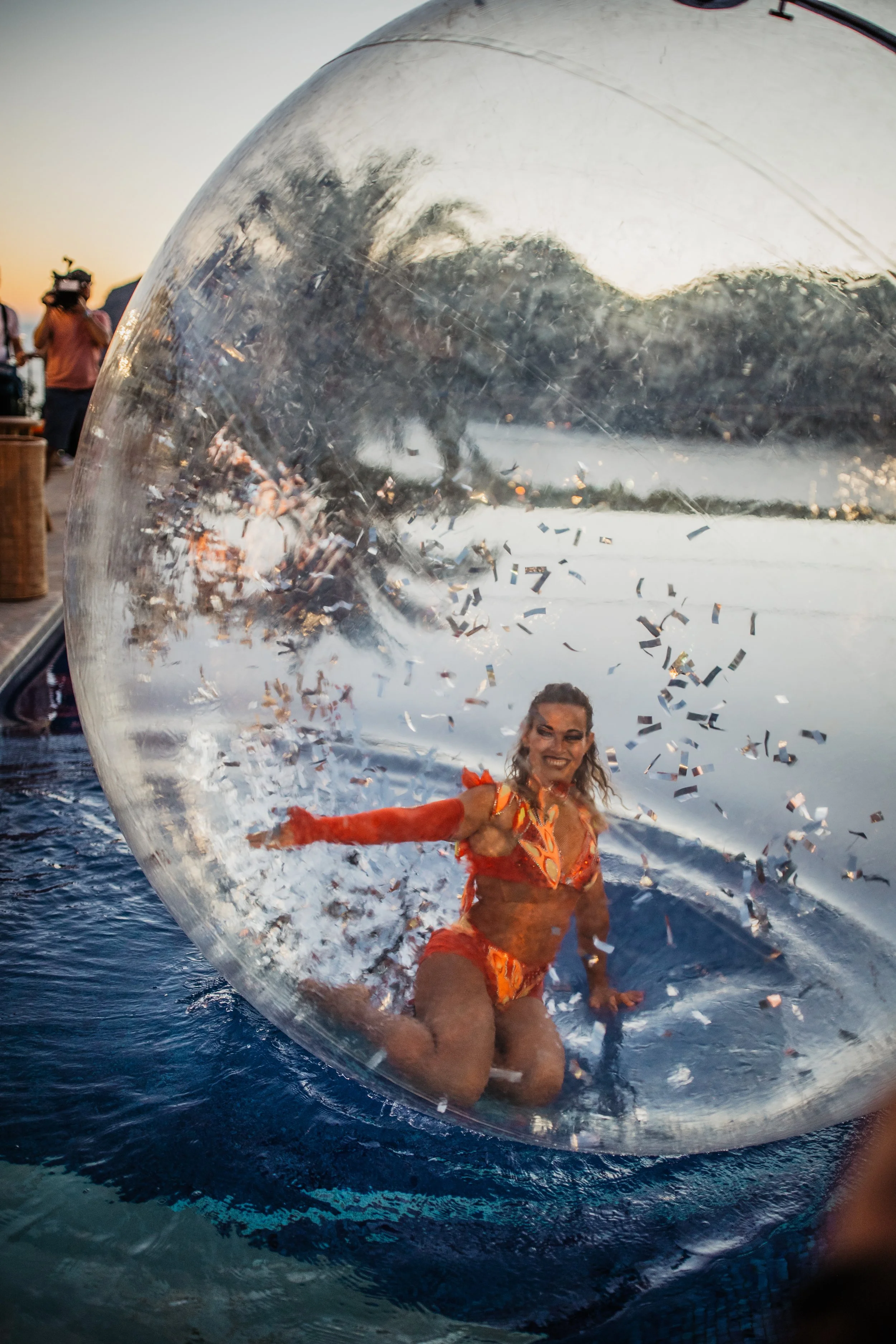 A woman in a colorful orange bikini posing inside a large, clear inflatable water ball on a body of water during sunset, with trees and people in the background.