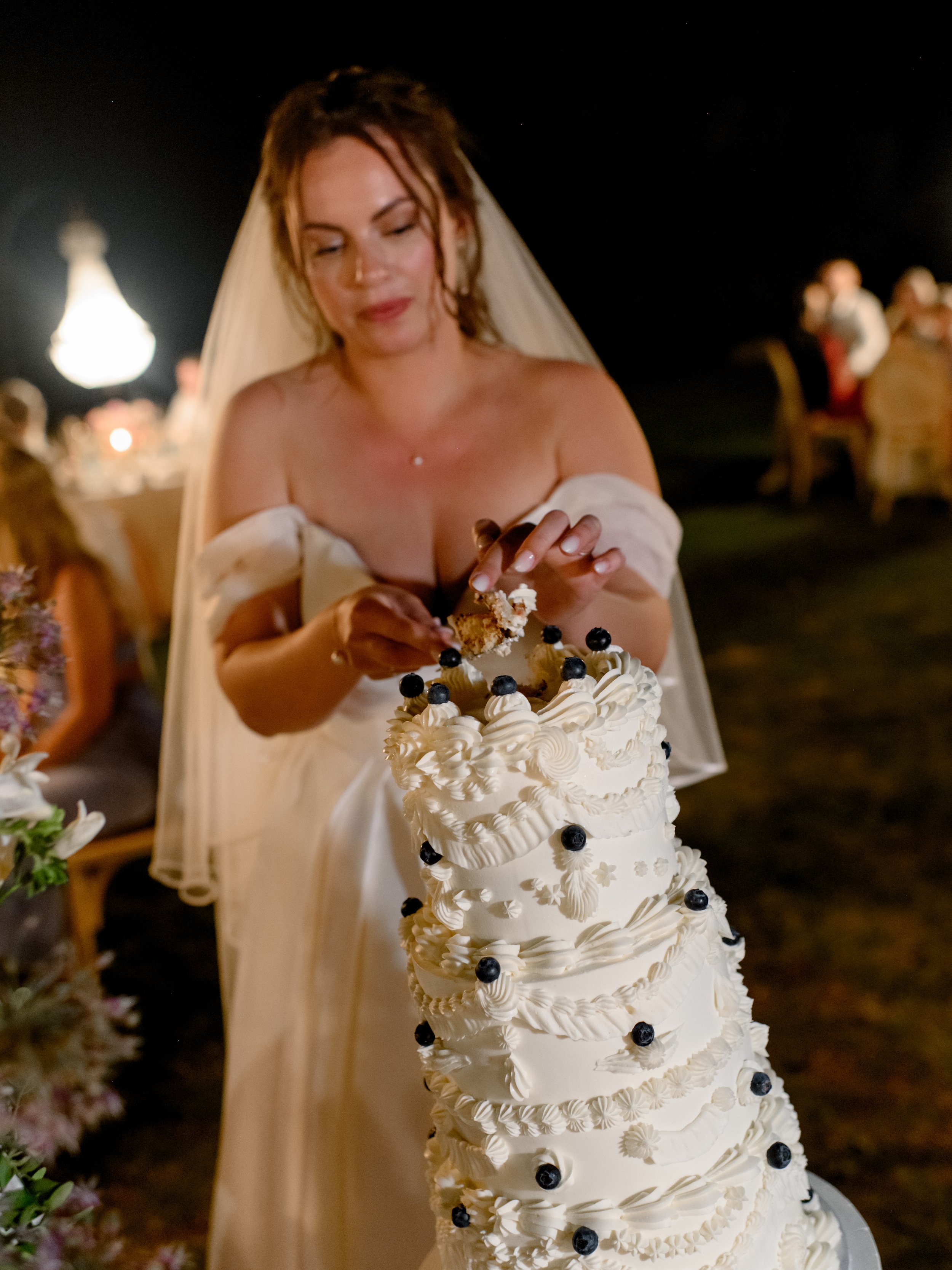 A bride wearing a white wedding dress and veil cuts into a tall, layered white wedding cake decorated with white icing swirls and blueberries at a nighttime outdoor reception.