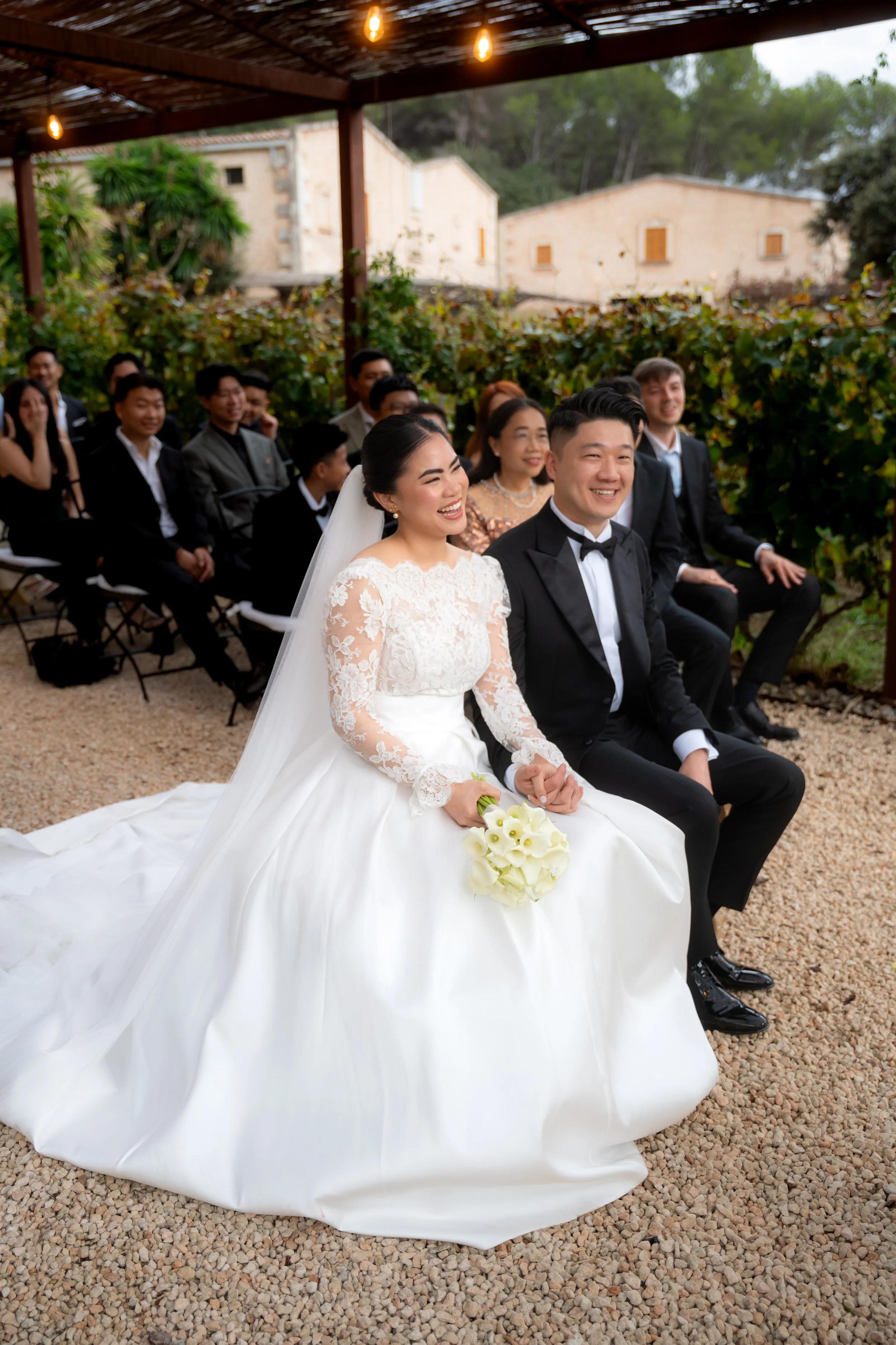 Bride and groom sitting outdoors at their wedding ceremony, surrounded by guests, with greenery and rustic buildings in the background.