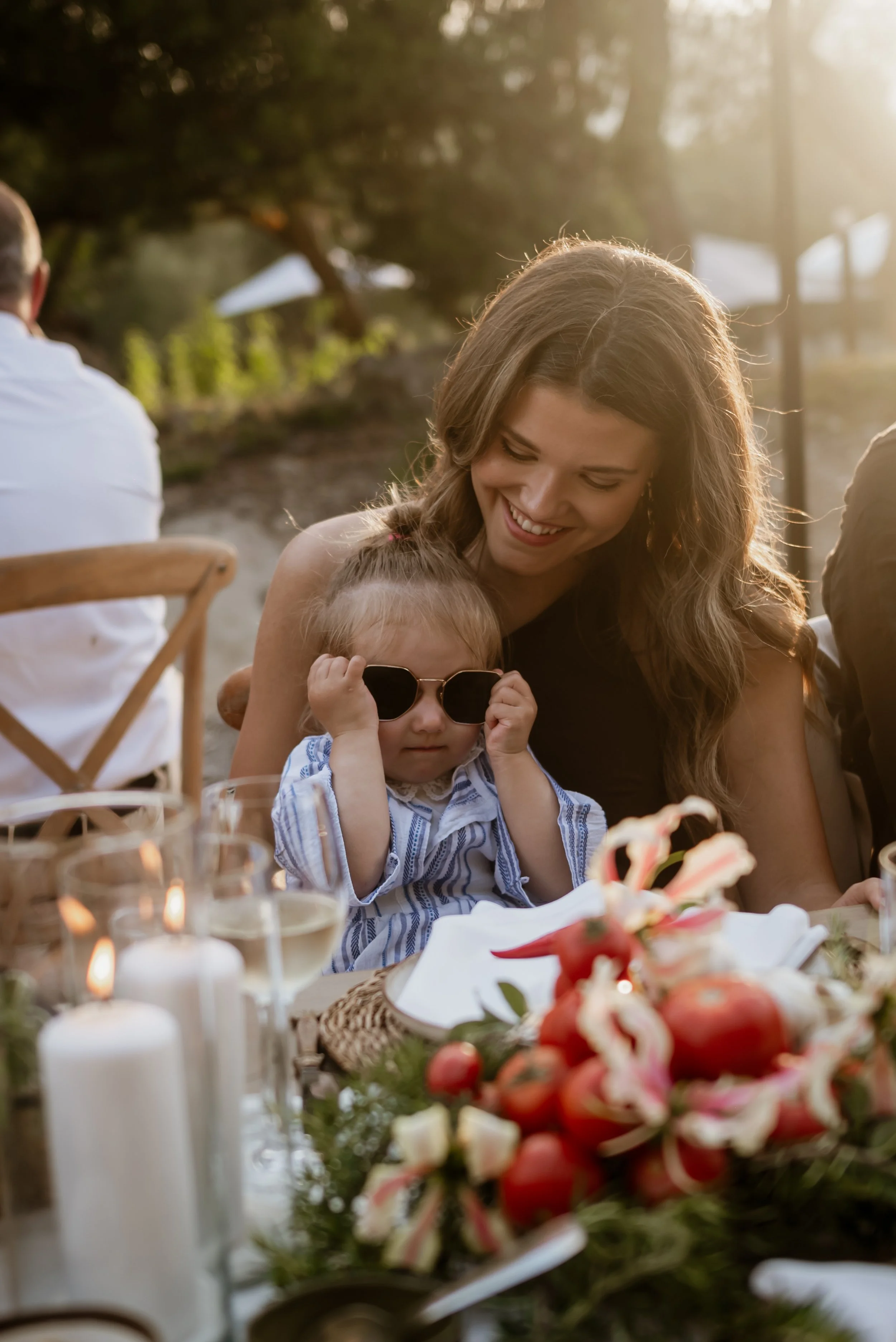 A young woman and a small girl wearing sunglasses sitting at a decorated outdoor table with candles and floral arrangements, smiling and enjoying a special occasion in the sunlight.