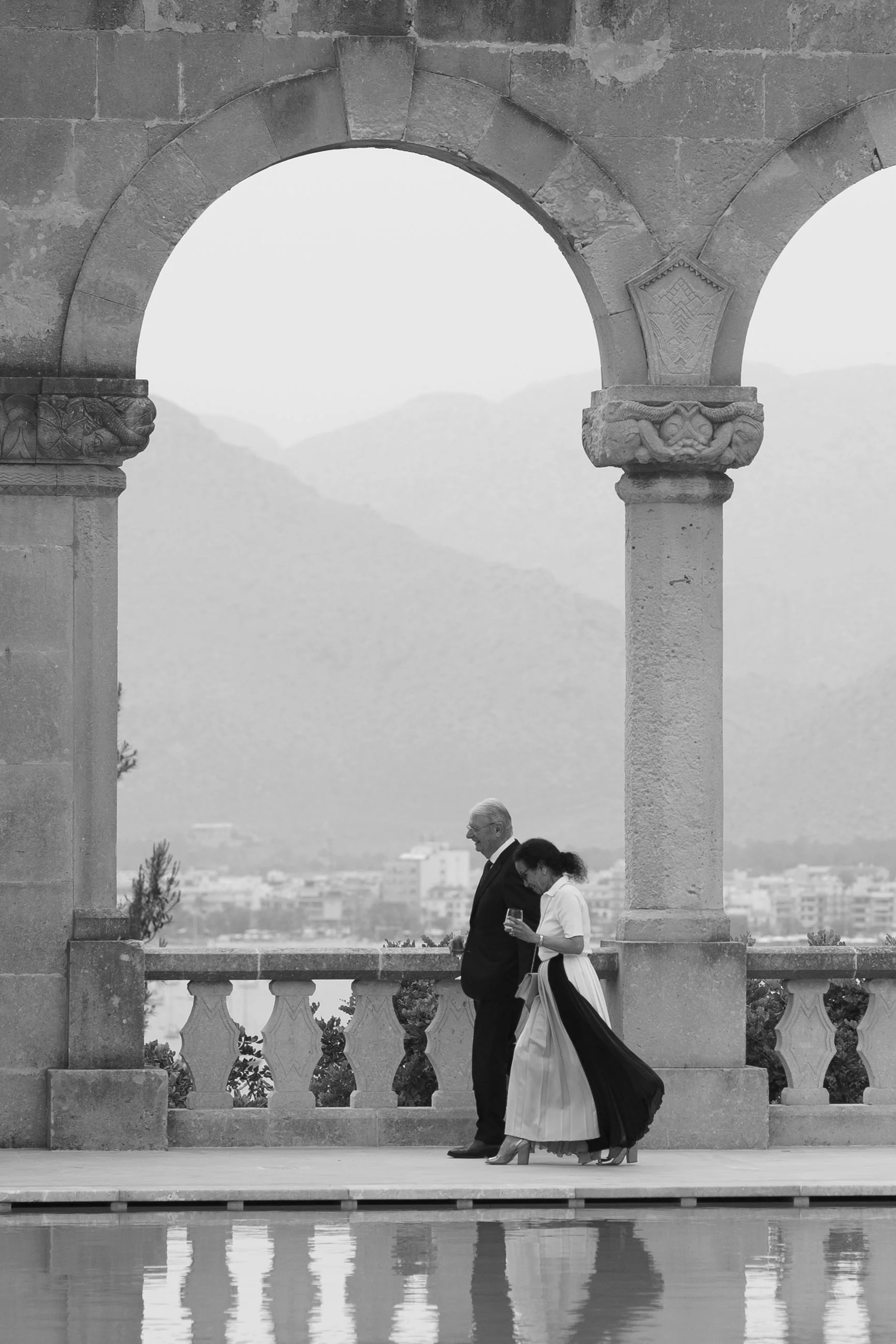 Two people, a man and a woman, walk together on a balcony with stone arches, overlooking a city and mountains in the background. The woman holds a glass of wine and wears a flowing dress, while the man is dressed in a suit.