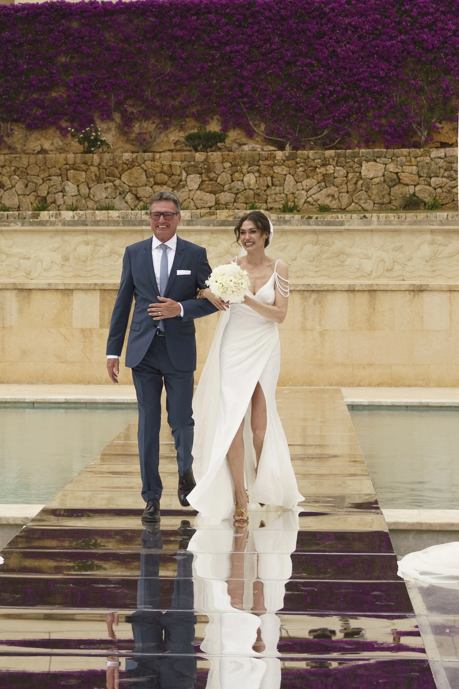 A bride and a man, possibly her father, walking down an aisle during a wedding ceremony. The bride is holding a bouquet of white flowers and is wearing a white wedding dress with a thigh-high slit and bead details on the shoulders. The man is wearing