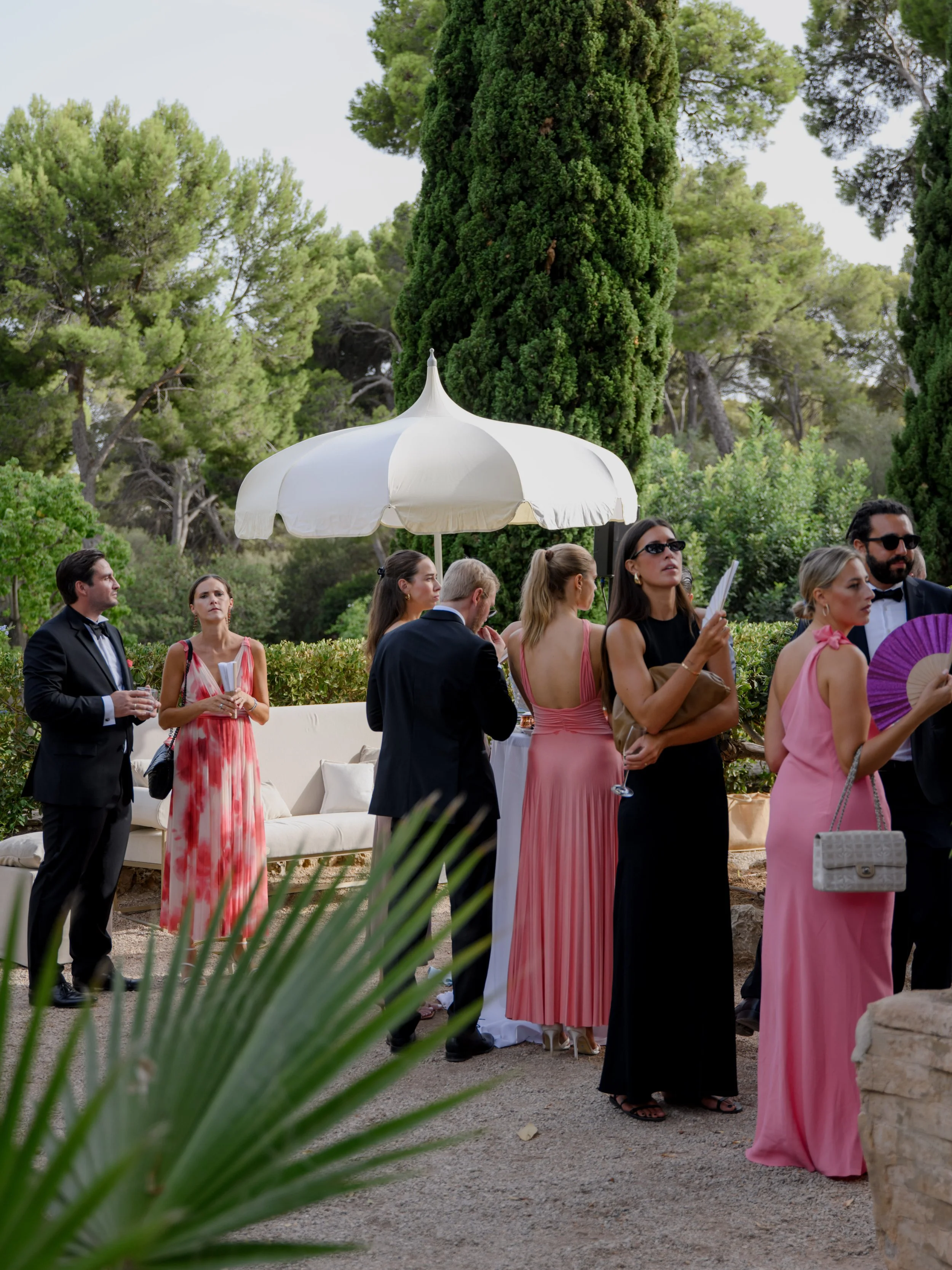 Group of elegantly dressed people at an outdoor event or party under an umbrella, surrounded by tall green trees and bushes.