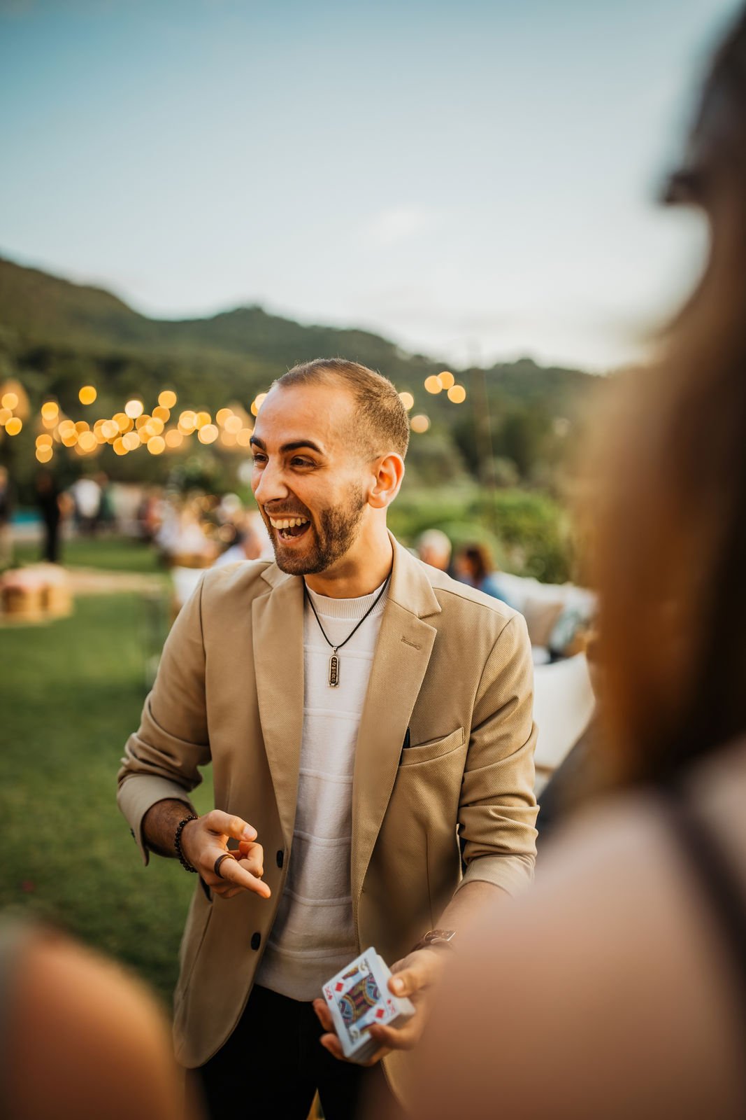 A man with a beard, wearing a beige blazer and a necklace, is smiling and engaging in a conversation at an outdoor event during the evening, with string lights and hills in the background.