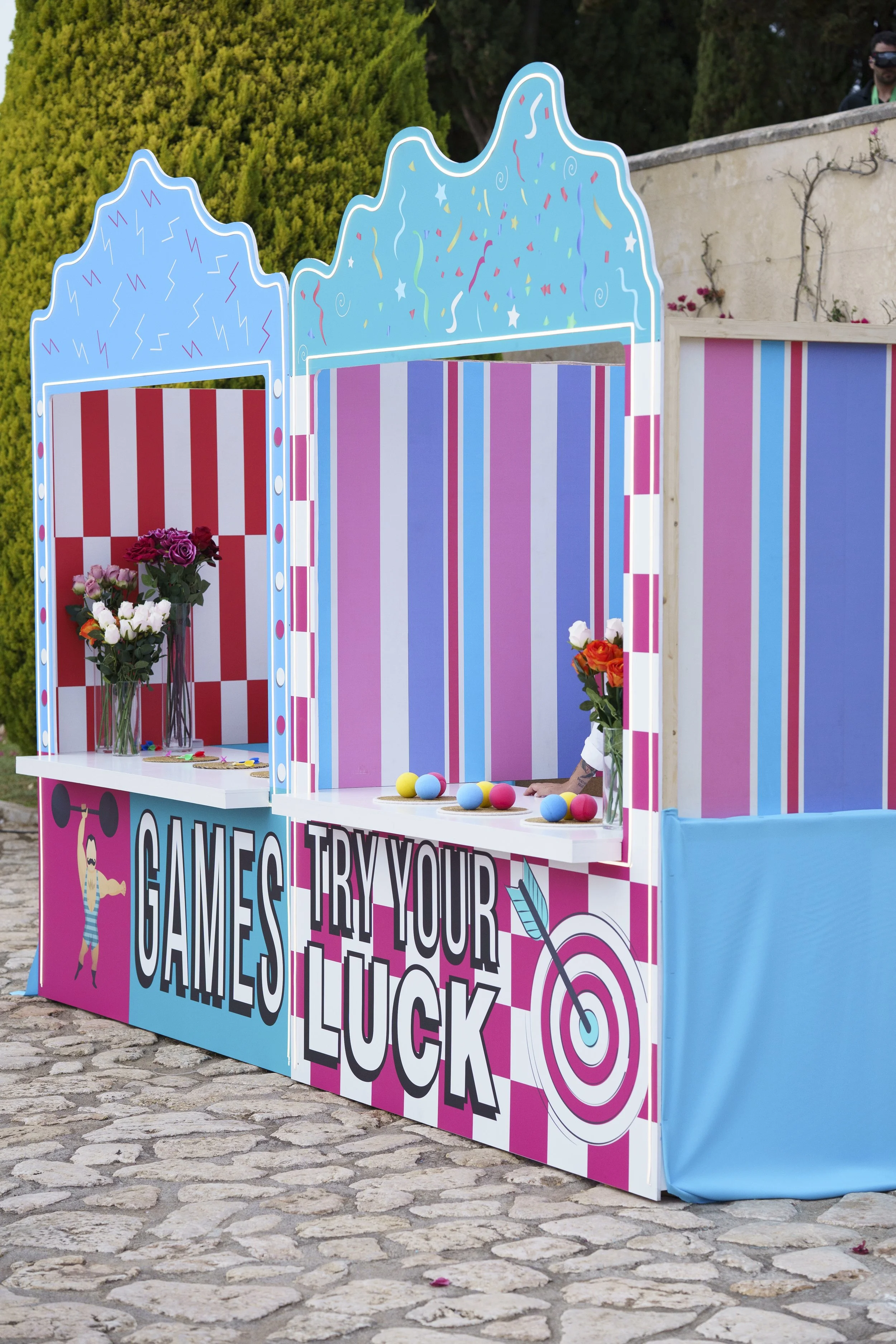 Colorful outdoor game booth with striped panels, featuring signs that say "GAMES" and "TRY YOUR LUCK," decorated with flowers and plastic balls for a carnival or festival.