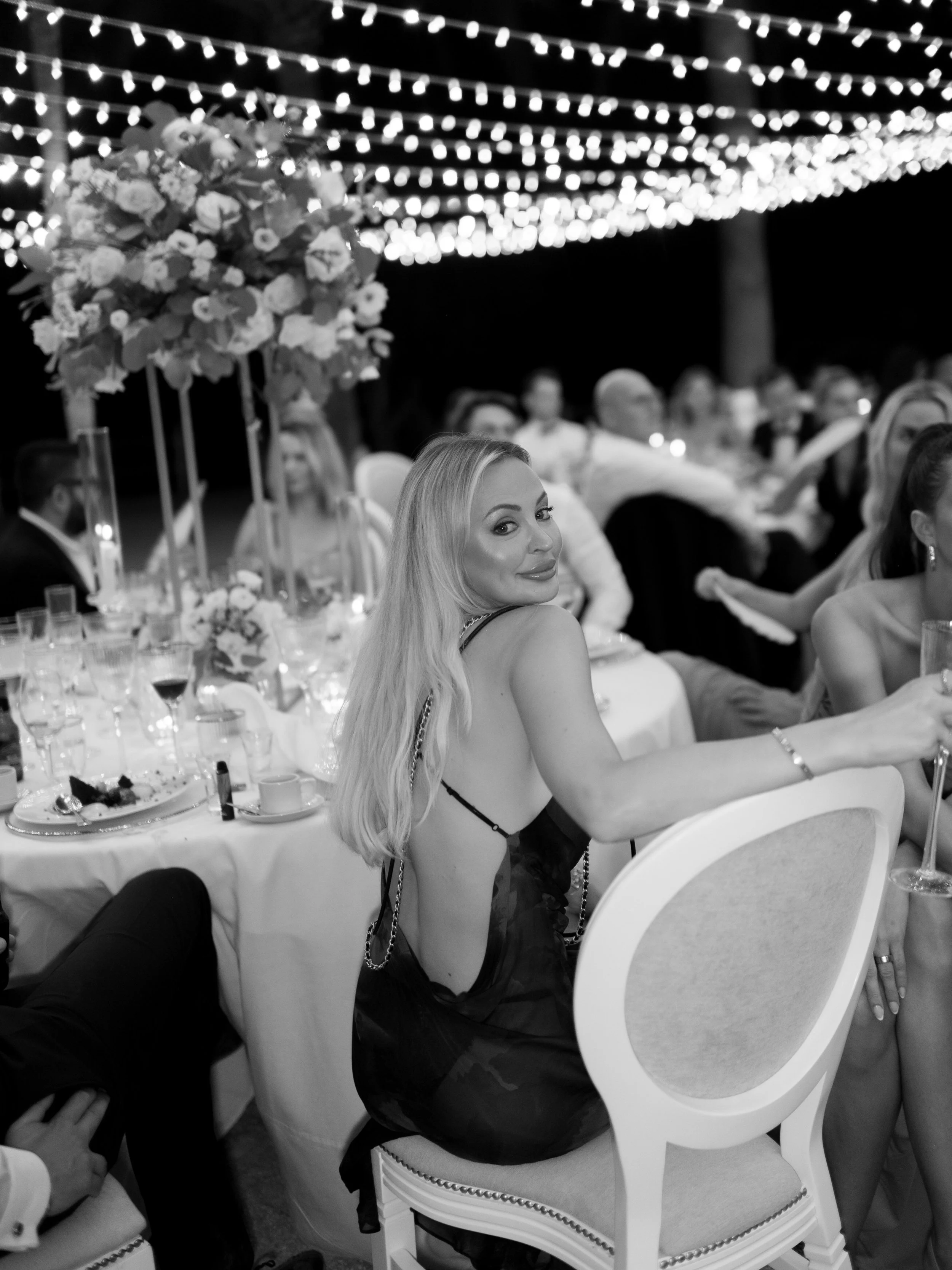 A woman with long blonde hair, wearing a black dress with open sides, sitting at a decorated table during a wedding or celebration, with string lights overhead.