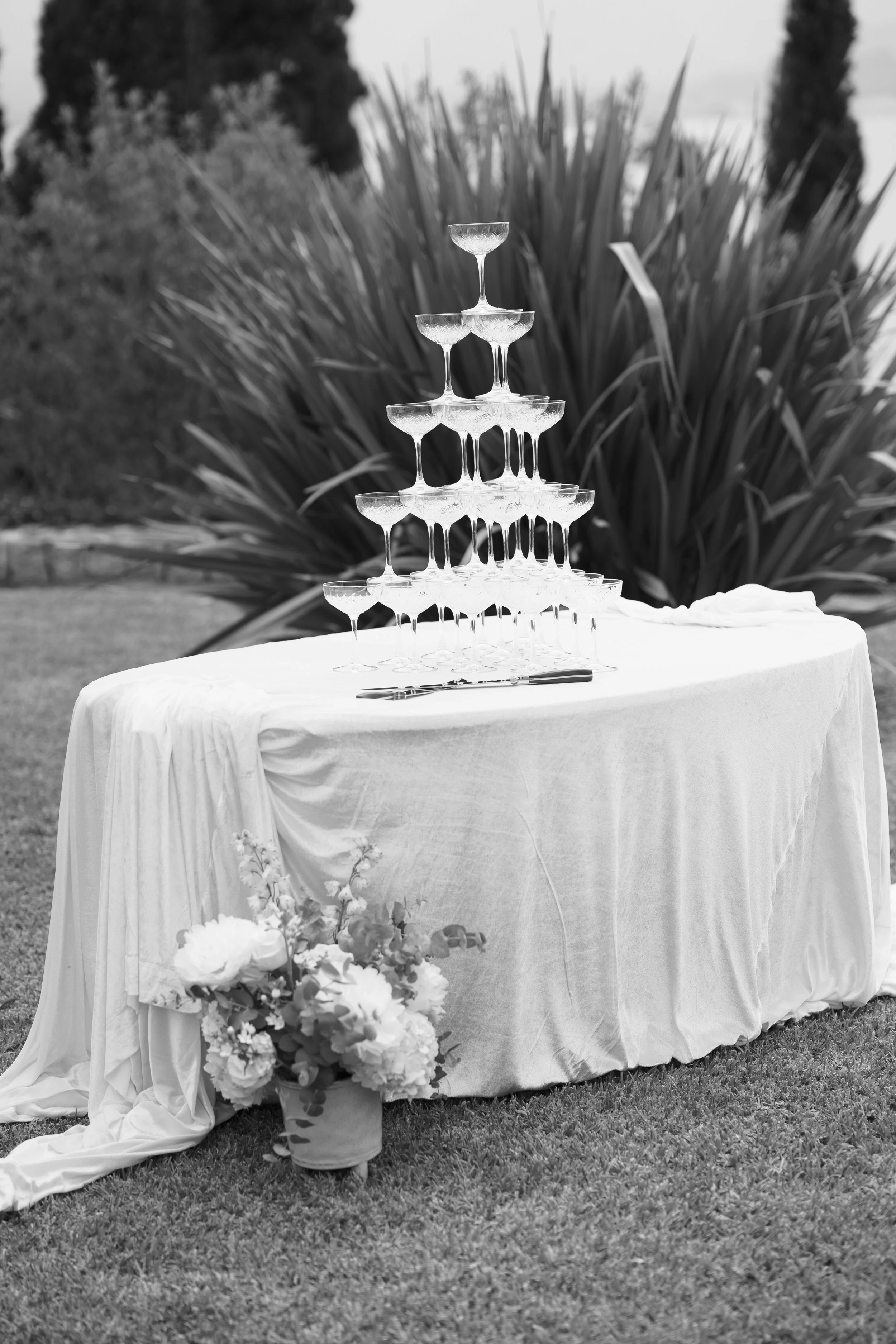A tiered display of champagne glasses arranged in a pyramid on a table with a white cloth, outdoors with surrounding plants and a flower arrangement in a pot.