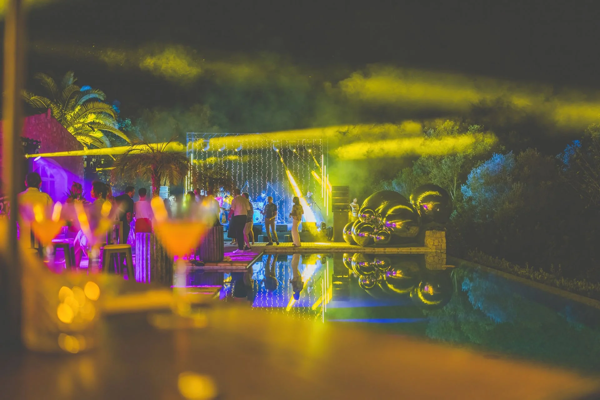 A nighttime outdoor party with colorful lighting, a stage with a band, balloons, and people dancing near a pool.