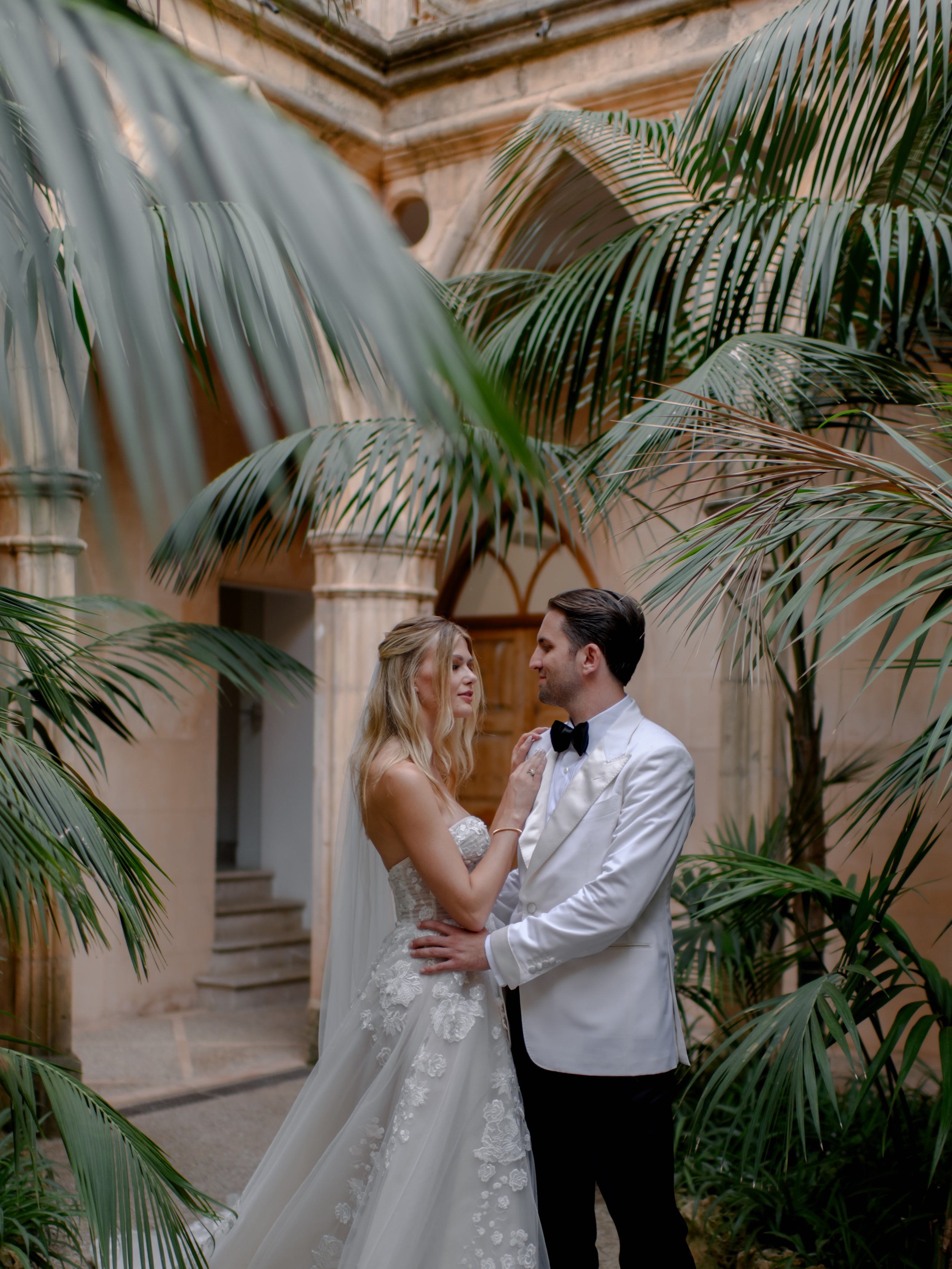 A bride and groom in wedding attire standing closely inside an elegant indoor garden with large green palm leaves and a stone architectural background.