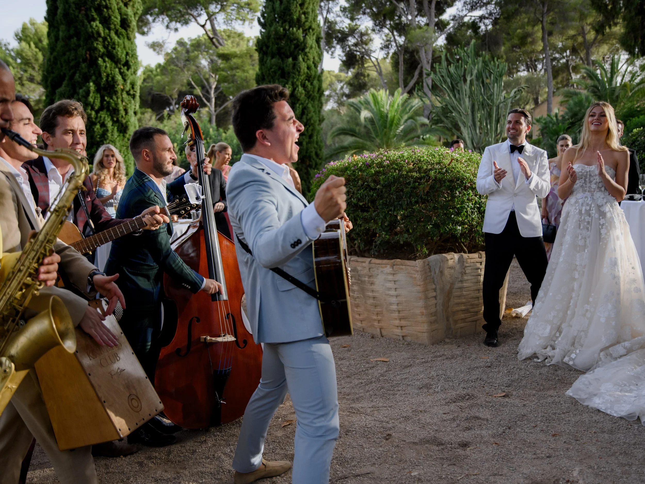 A wedding celebration with a live band performing outdoors, including a singer with a guitar, and a bride and groom clapping and smiling.
