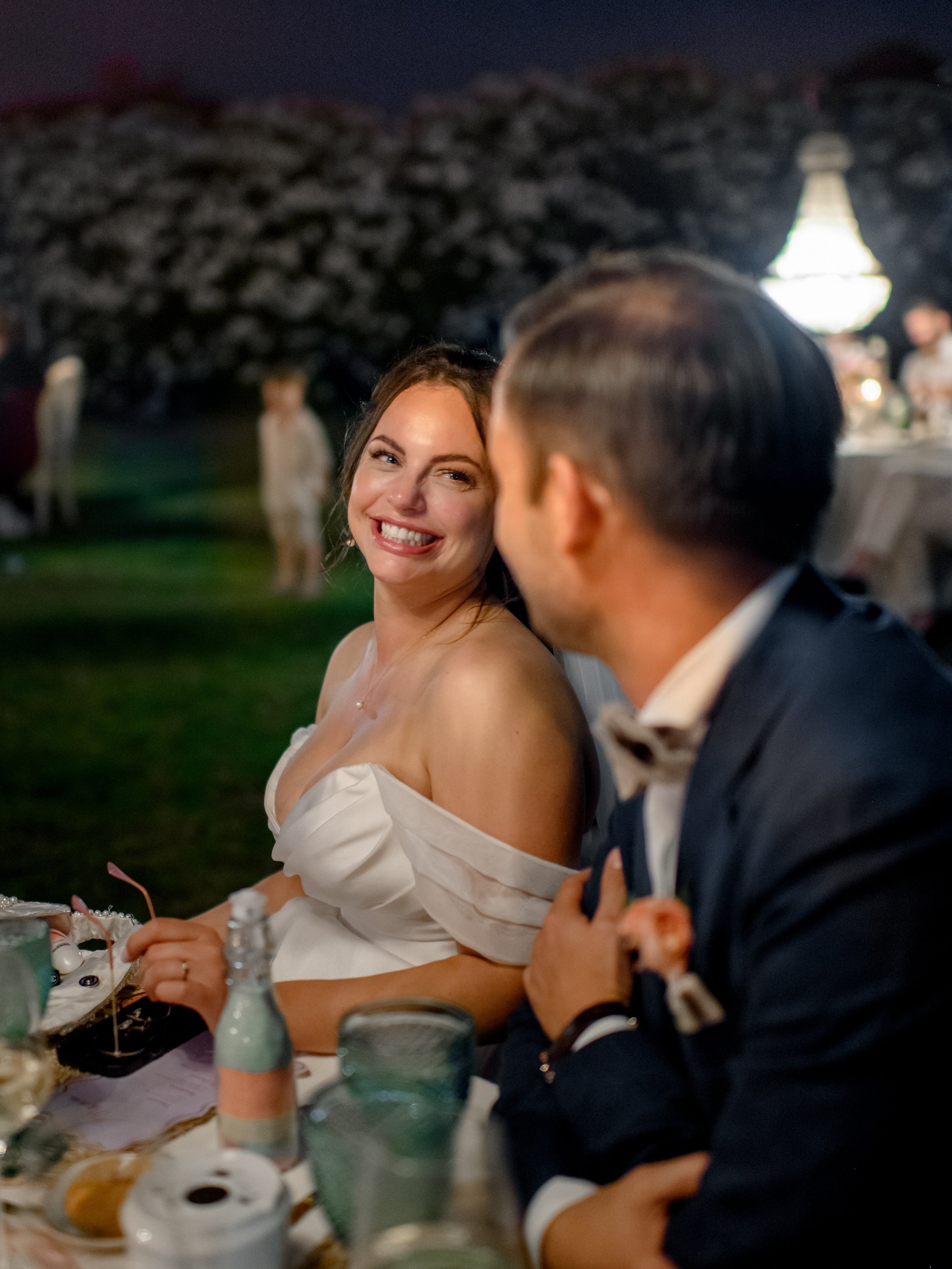 A woman in a white off-shoulder dress smiling at a man in a tuxedo at an outdoor evening event with a lit lantern in the background.