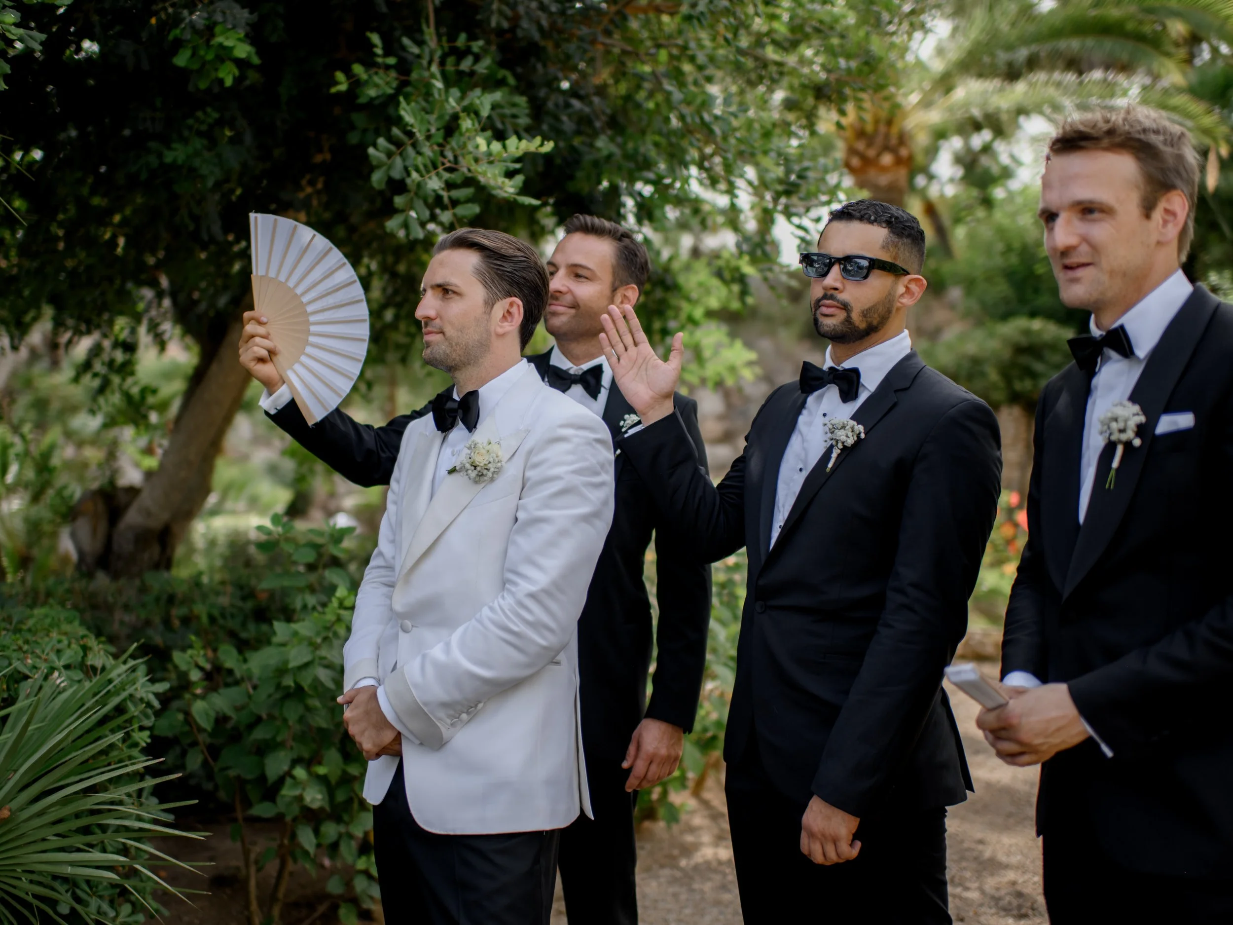 Four men dressed in black tuxedos and a man in a white tuxedo with boutonnières, standing outdoors among green trees, some with their eyes closed, one holding a fan, one raising his hand, and another holding a paper, during a formal event.