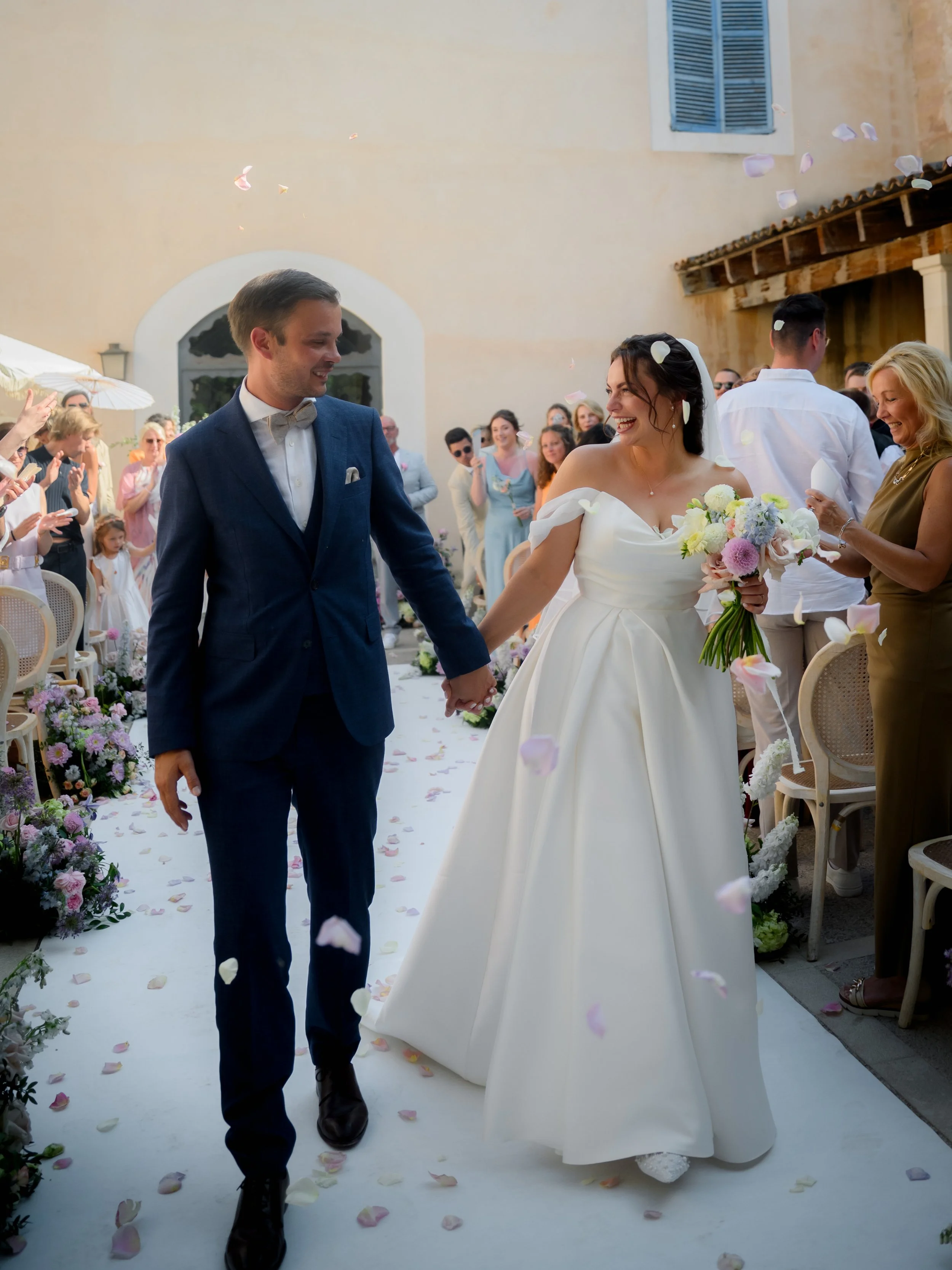 A bride and groom walking hand in hand down the aisle after their wedding ceremony, surrounded by guests and falling flower petals.