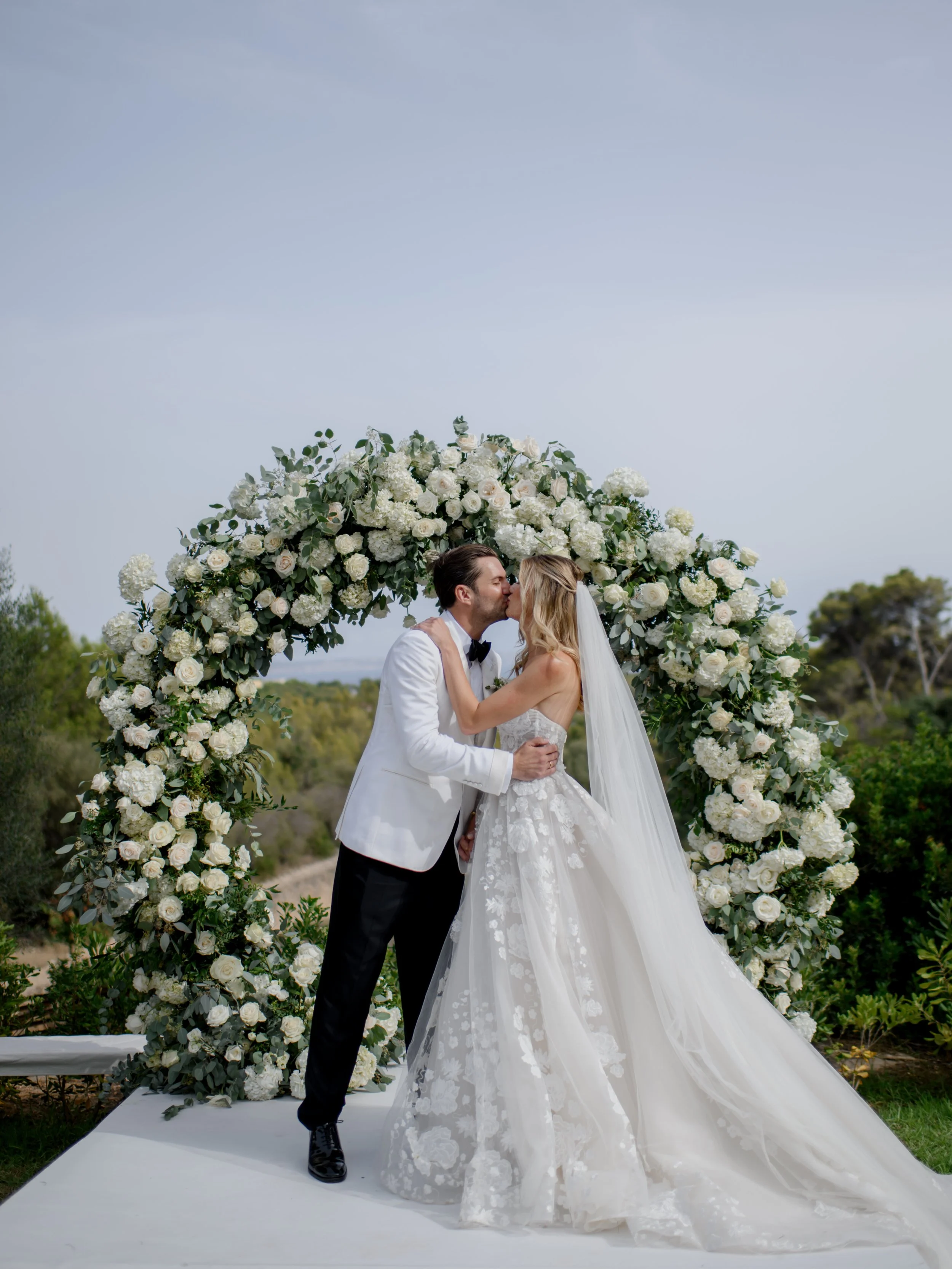 A bride and groom kiss under a large floral arch with white roses and greenery, outdoors on a clear day.