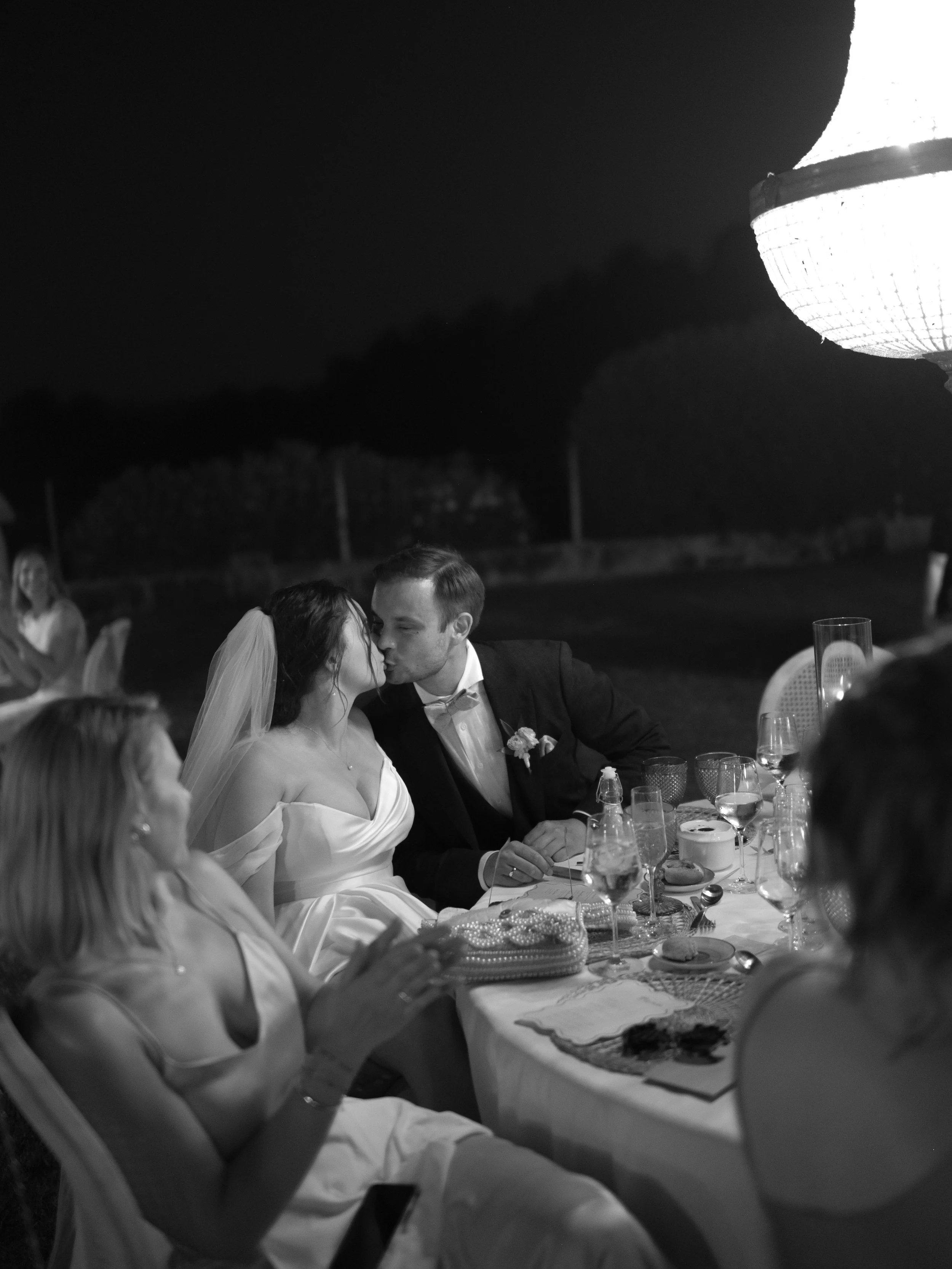 A black-and-white photo of a wedding reception outdoors at night, showing a bride and groom kissing at a decorated table. The bride is wearing a wedding dress with a veil, and the groom is in a tuxedo. Guests are seated around the table with glasses 